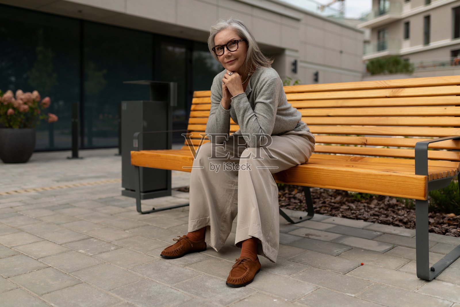 白天，一位年长的女士舒适地坐在现代户外环境的木制长凳上。照片摄影图片