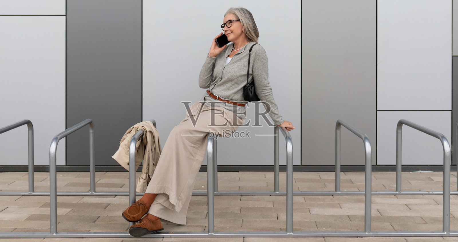 一位年长的女性坐在建筑外的栏杆上，悠闲地用手机交谈。照片摄影图片