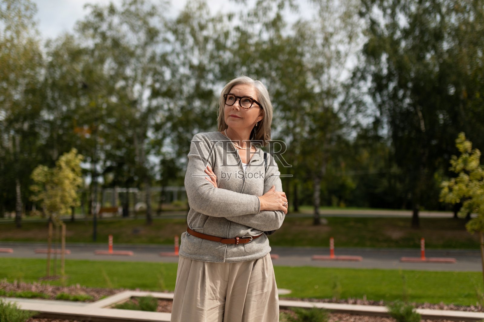一位年长的女士自信地站在公园里，在绿树和景观区域的环绕下，享受着晴朗的户外时光。照片摄影图片