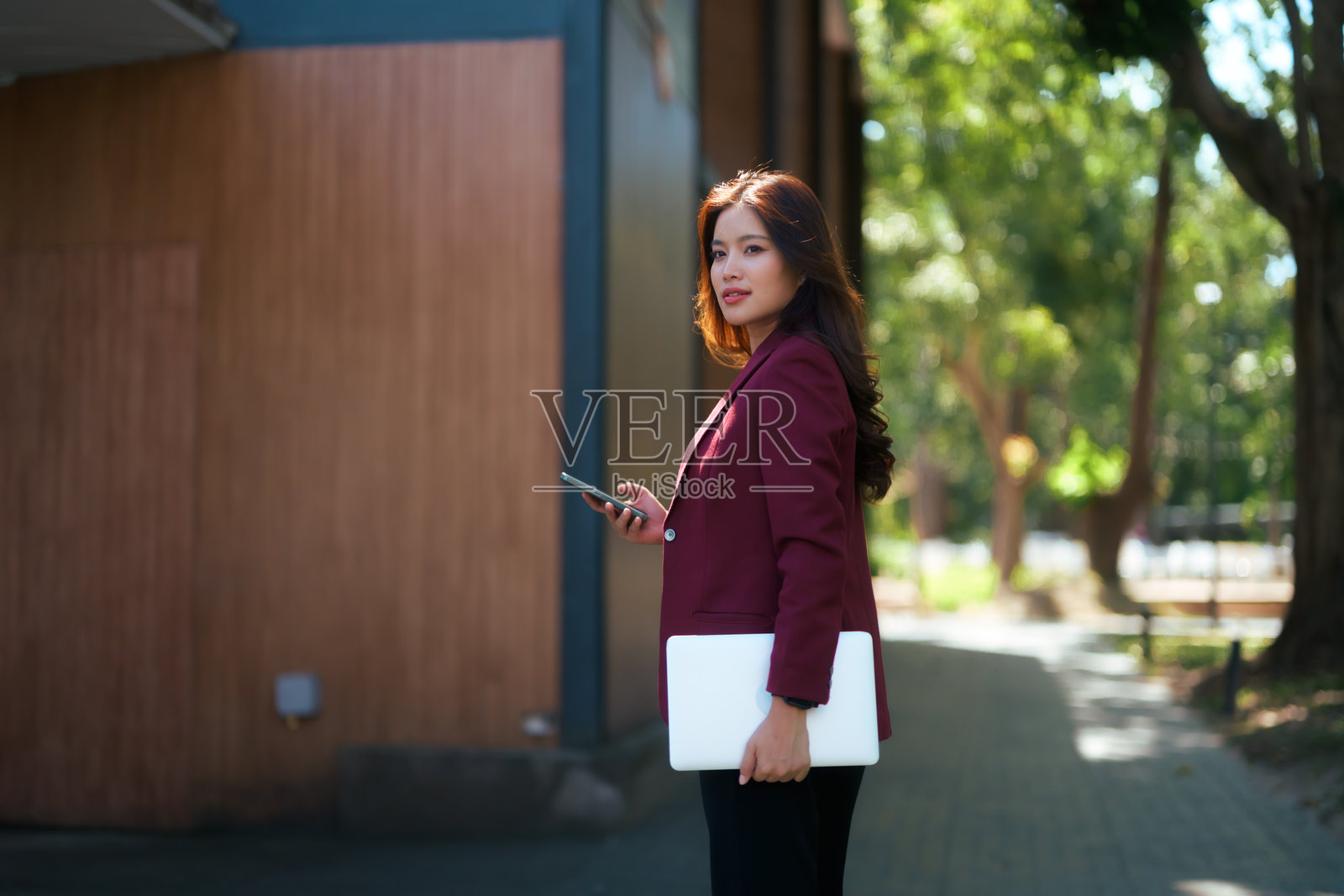 一位亚洲女商人在阳光明媚的日子里，自信地走在公园里，手里拿着笔记本电脑和智能手机，同时回头看向后方。照片摄影图片