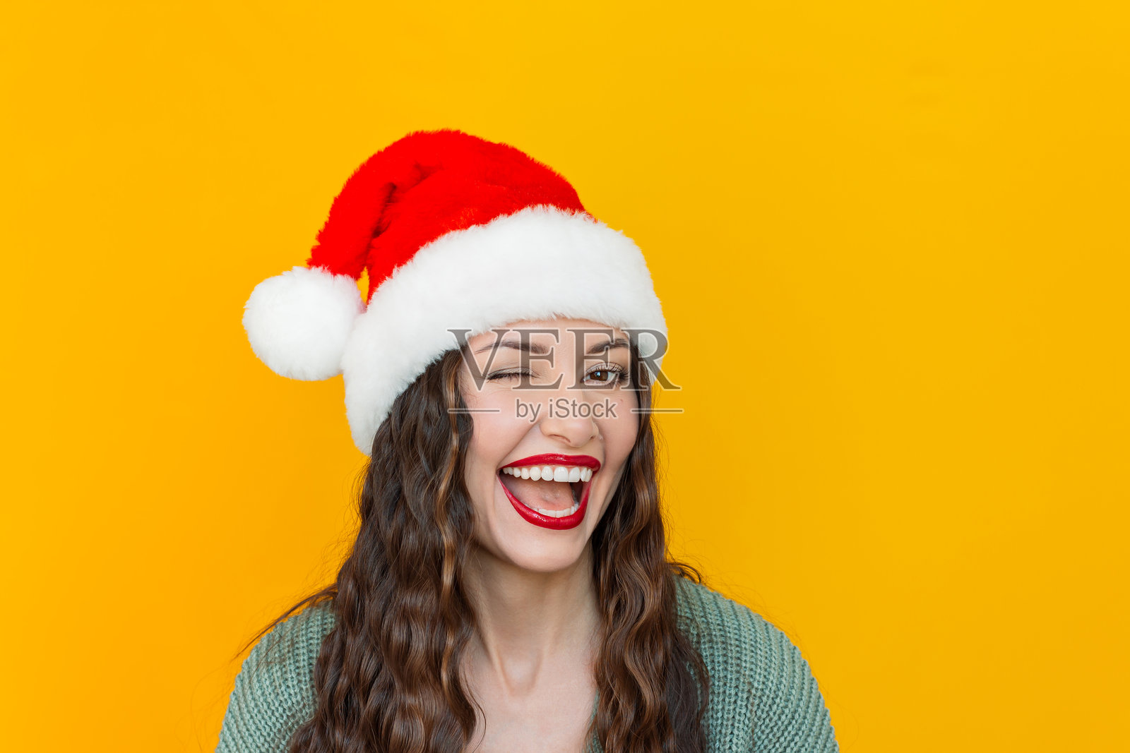 年轻女子戴着圣诞帽，在黄色背景下对着镜头眨眼微笑。照片摄影图片