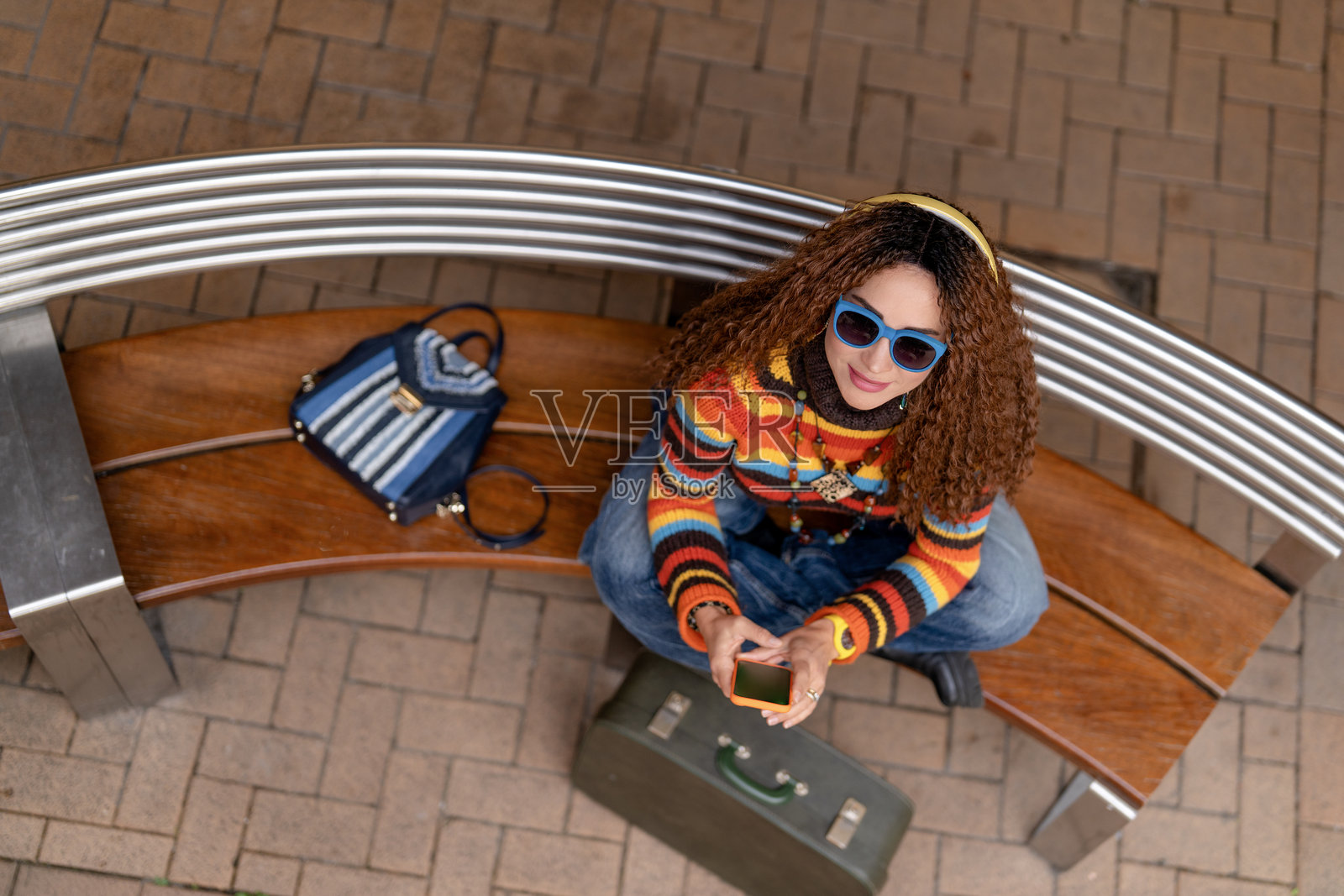 年轻女子白天在户外长凳上，穿着亮色毛衣，戴着时尚太阳镜，享受着她的智能手机。照片摄影图片