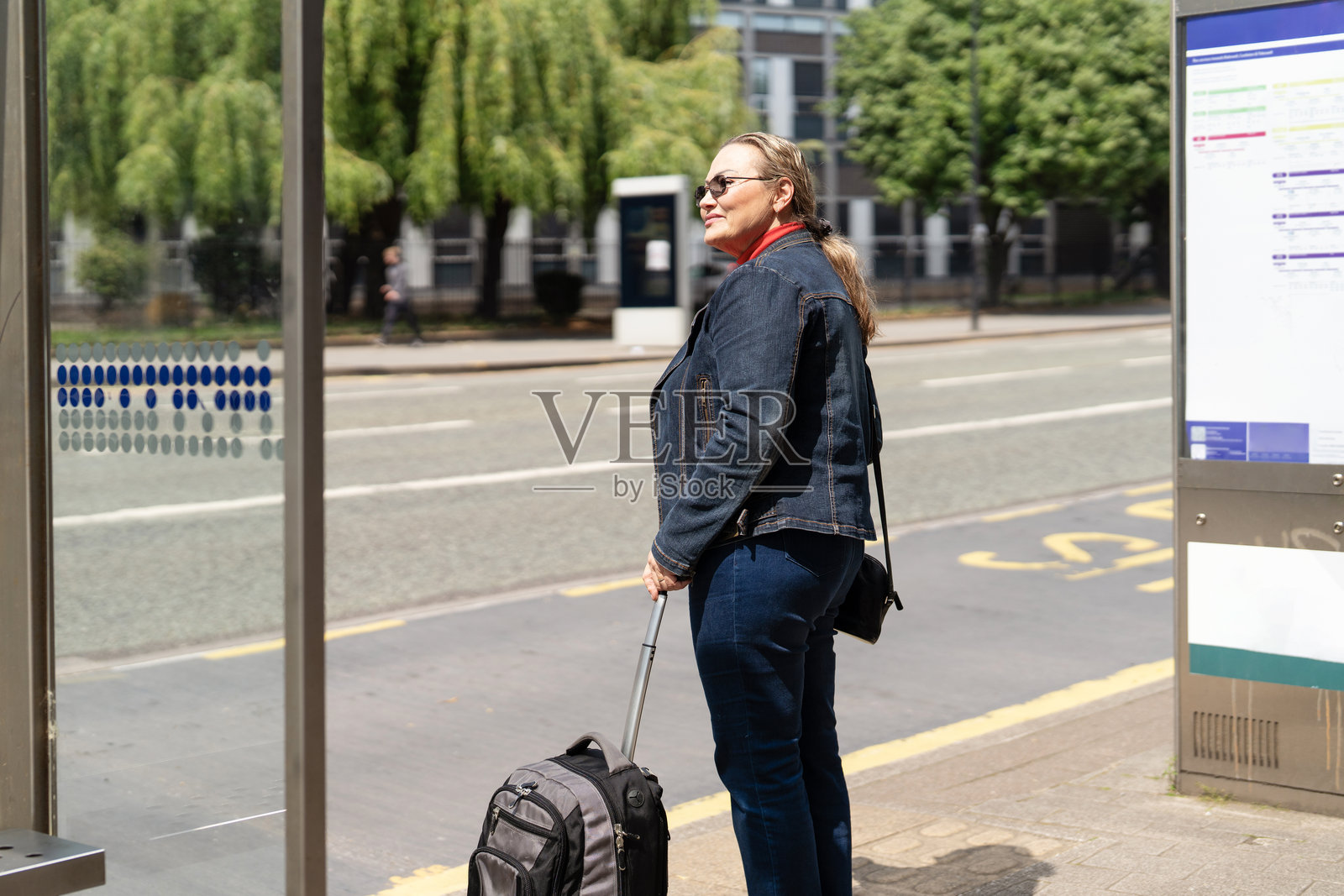 阳光明媚的城市里，一位女士带着行李在公交车站等车。照片摄影图片