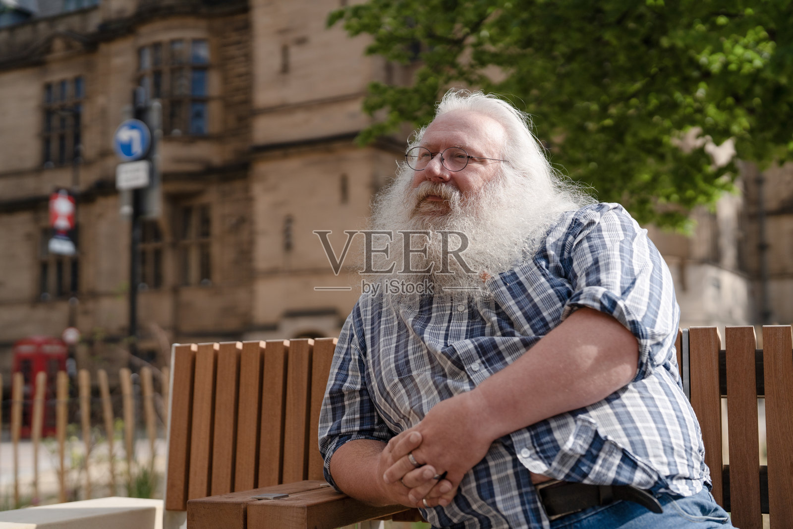 阳光明媚的日子里，一位长着长胡子的男士坐在城市街道历史建筑外的长椅上。照片摄影图片