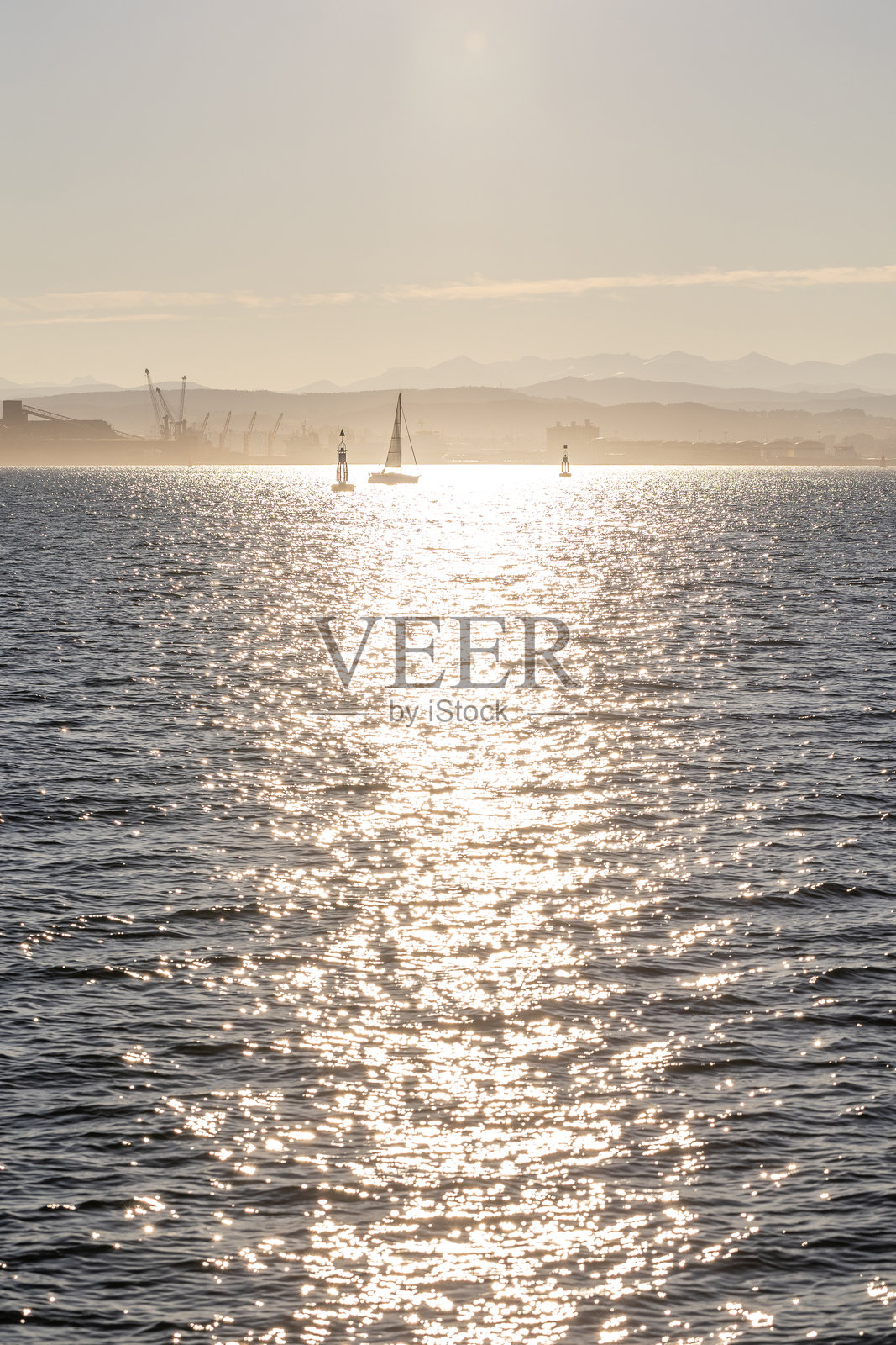 在阳光明媚的日子里，一艘孤独的帆船在桑坦德湾的蓝色水面上航行，远处可以看到海岸线和山脉。照片摄影图片