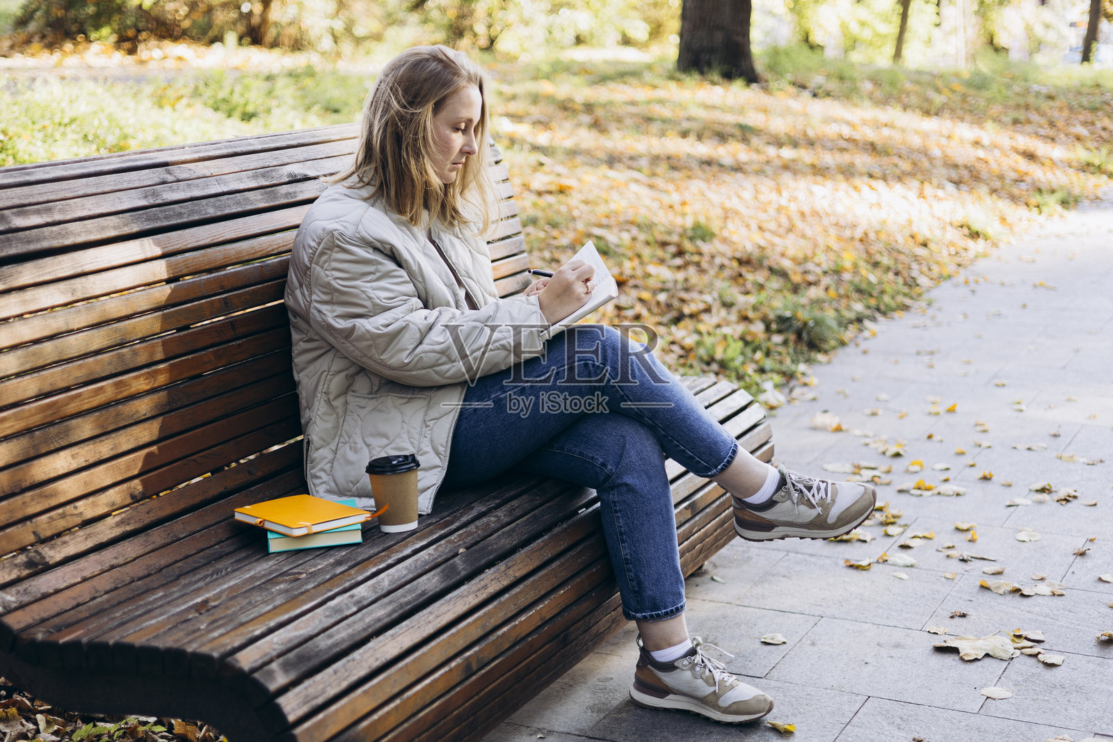 年轻女子在秋季公园的长椅上用笔记本写字。照片摄影图片