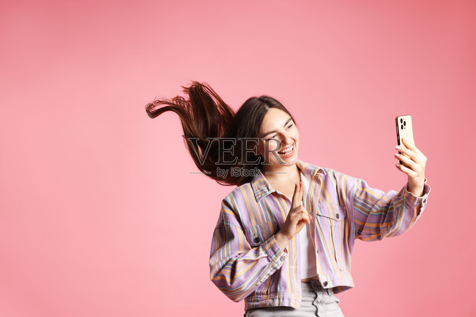 穿着夹克的长发女子在粉色背景前用智能手机自拍，并比出胜利手势，留有文字空间。照片摄影图片