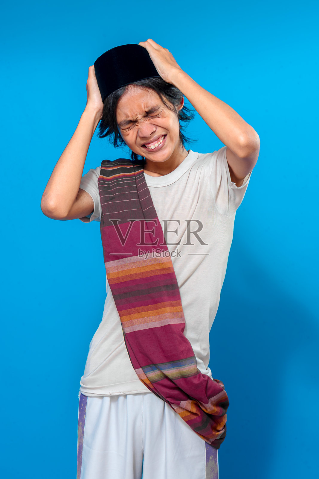 一名年轻的印度尼西亚男子身着传统服饰，戴着佩奇帽，裹着纱笼，痛苦地皱着眉挠头，在蓝色背景下表达着沮丧或困惑。照片摄影图片
