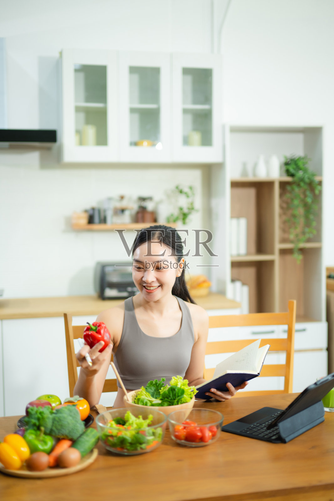 身着健身服的亚洲女性在厨房餐桌旁使用平板电脑，同时享用健康食品。她手中拿着番茄，并在笔记本上书写。主题是清洁饮食和远程健康生活方式。照片摄影图片