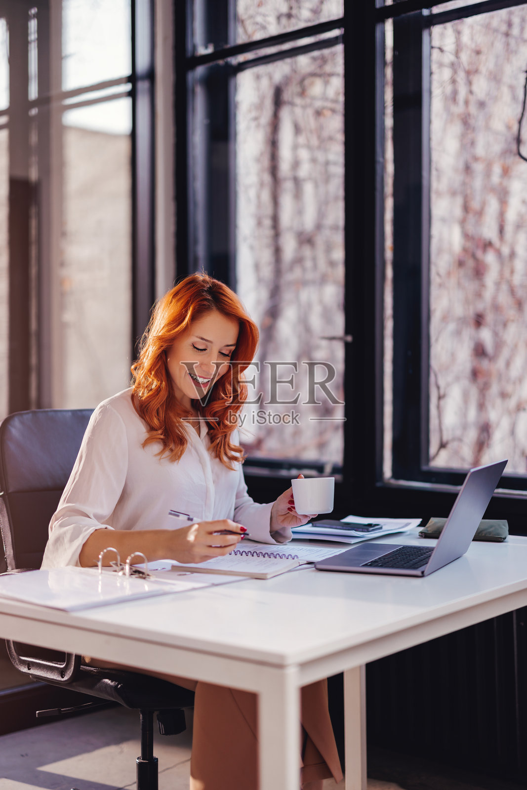 一位身着正装、面带微笑的快乐女商人在现代办公室的办公桌前，在笔记本电脑前处理文件。照片摄影图片