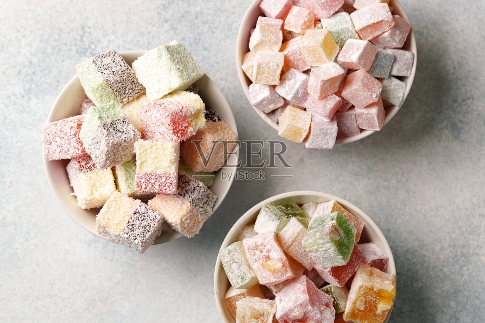 三种碗里摆放着色彩缤纷的土耳其软糖，表面撒着糖粉，置于浅色背景上照片摄影图片