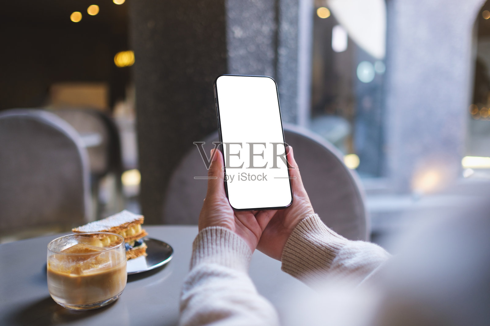 咖啡馆里，女子手持并使用手机，背景为空白的桌面屏幕的样机图照片摄影图片