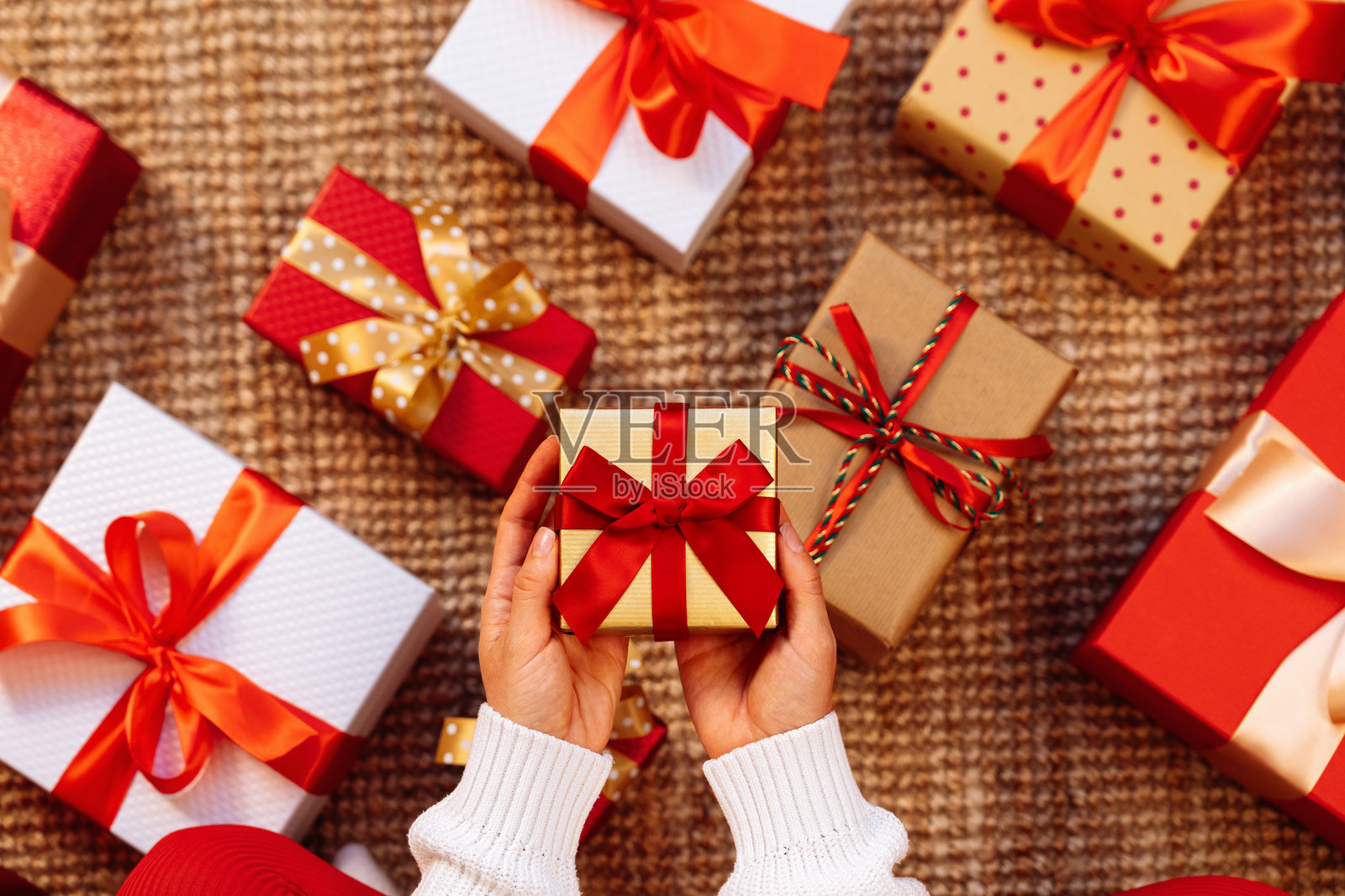手中拿着一个带红色丝带和蝴蝶结的小礼物，礼物上方是地板上的礼品盒。照片摄影图片