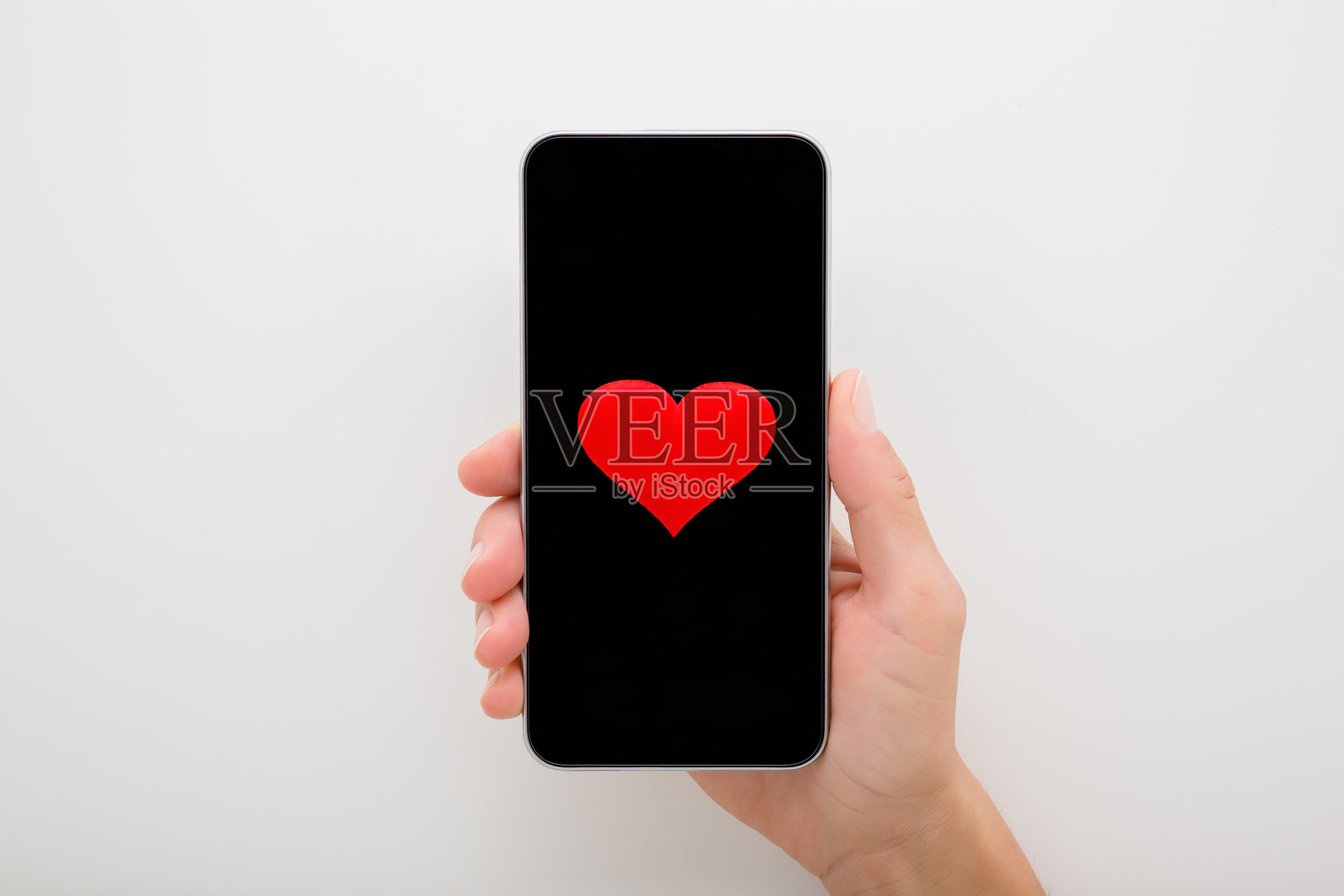 年轻女性手持智能手机，深黑色触摸屏，浅灰色桌面上有一个红心形状。爱意概念。在线约会。特写。俯视角度。照片摄影图片