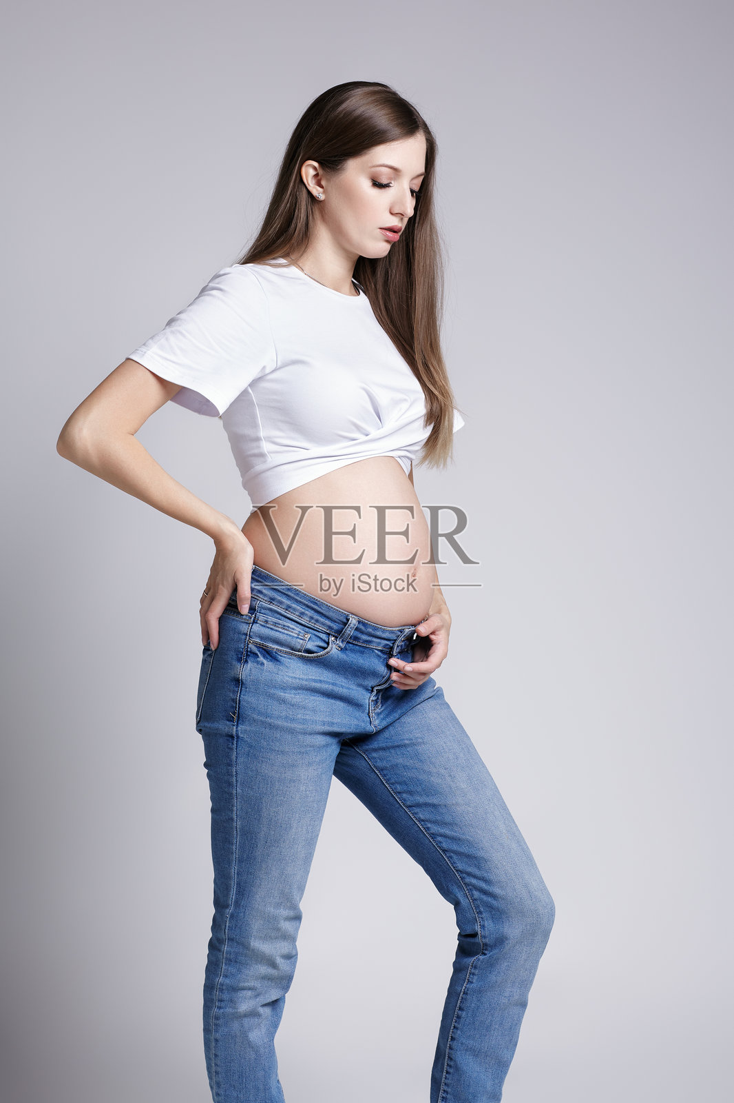 年轻漂亮的孕妇，穿着白色T恤和牛仔裤，看着自己的孕肚露出，怀孕5个月。照片摄影图片