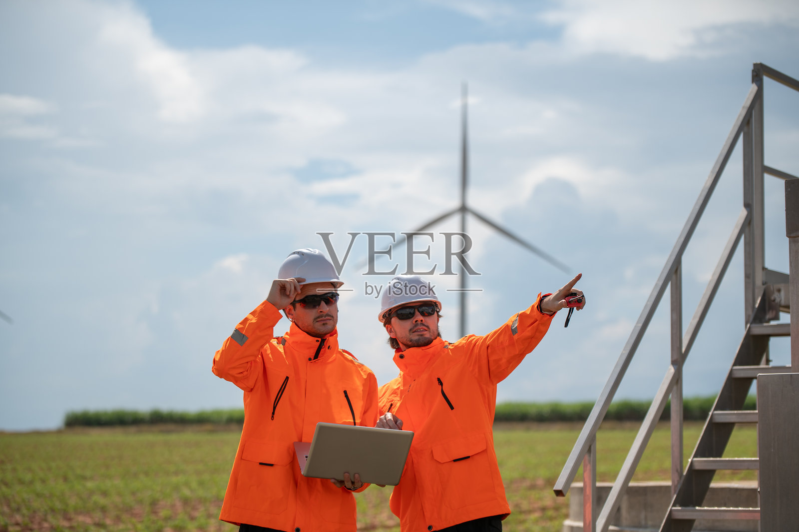 工程师们戴着安全帽，在风力发电场检查风力涡轮机，并拿着笔记本电脑工作。照片摄影图片