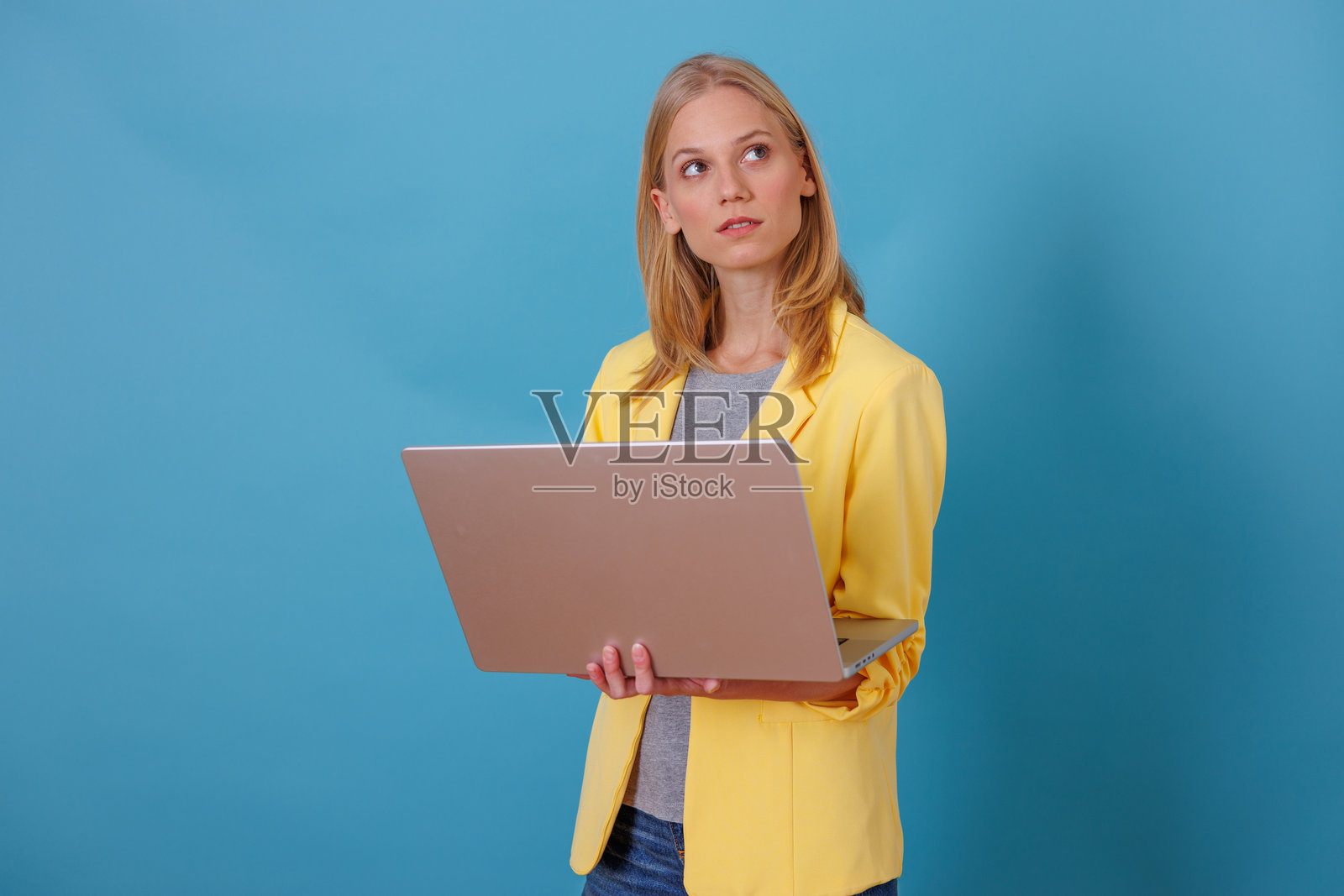 身穿时尚黄色西装外套的年轻女子，手持笔记本电脑，背景为蓝色，代表现代创意照片摄影图片