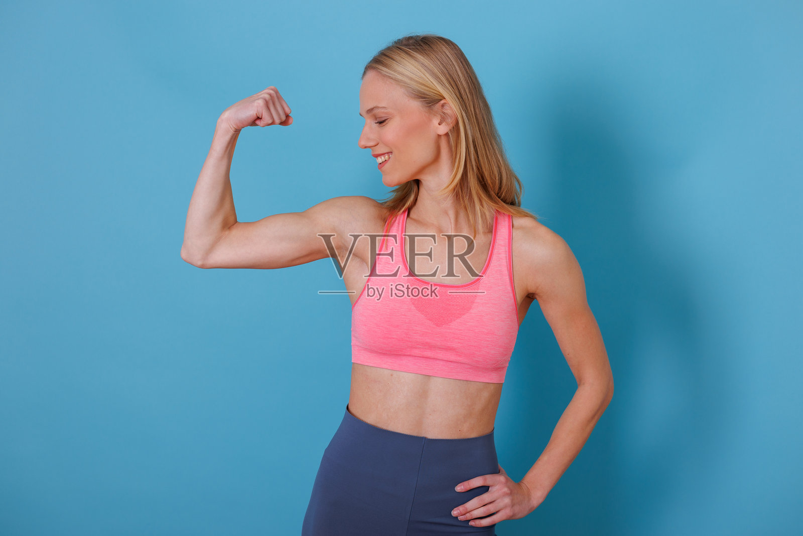一名年轻女子穿着粉色运动内衣，在纯蓝色背景前自信地展示肱二头肌并微笑，这是腰部以上的影棚拍摄。照片摄影图片