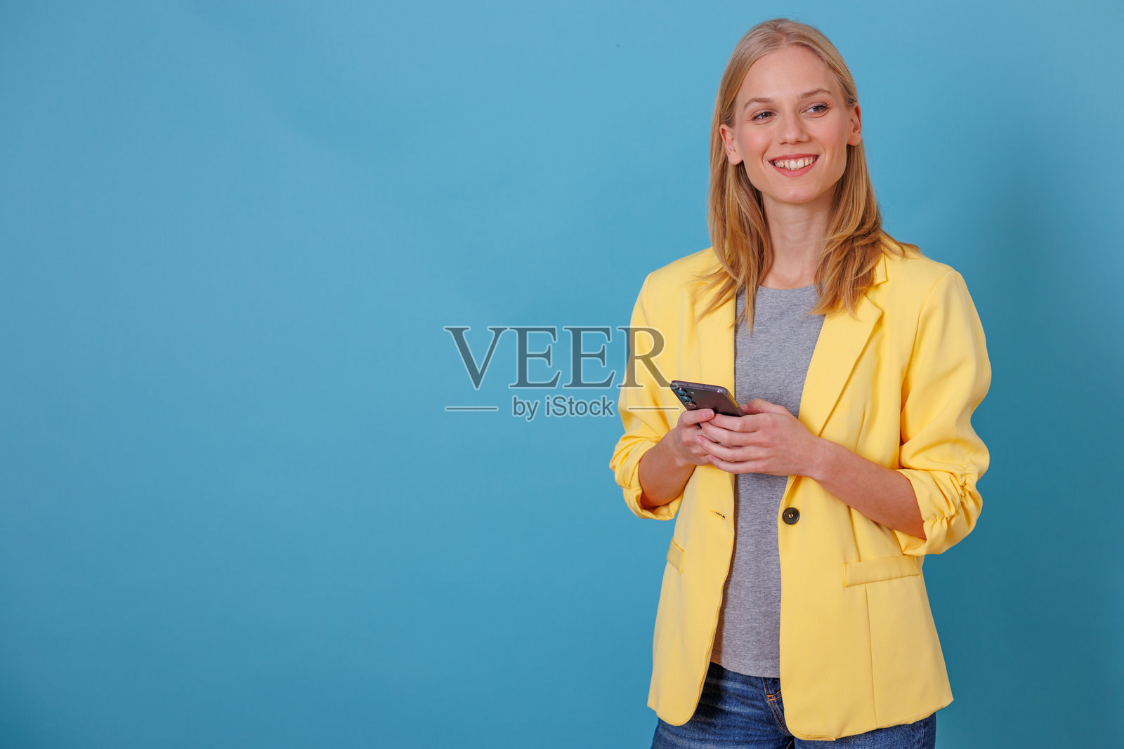 年轻女子对着蓝色背景微笑，使用智能手机发短信和浏览网页，留有复制空间照片摄影图片