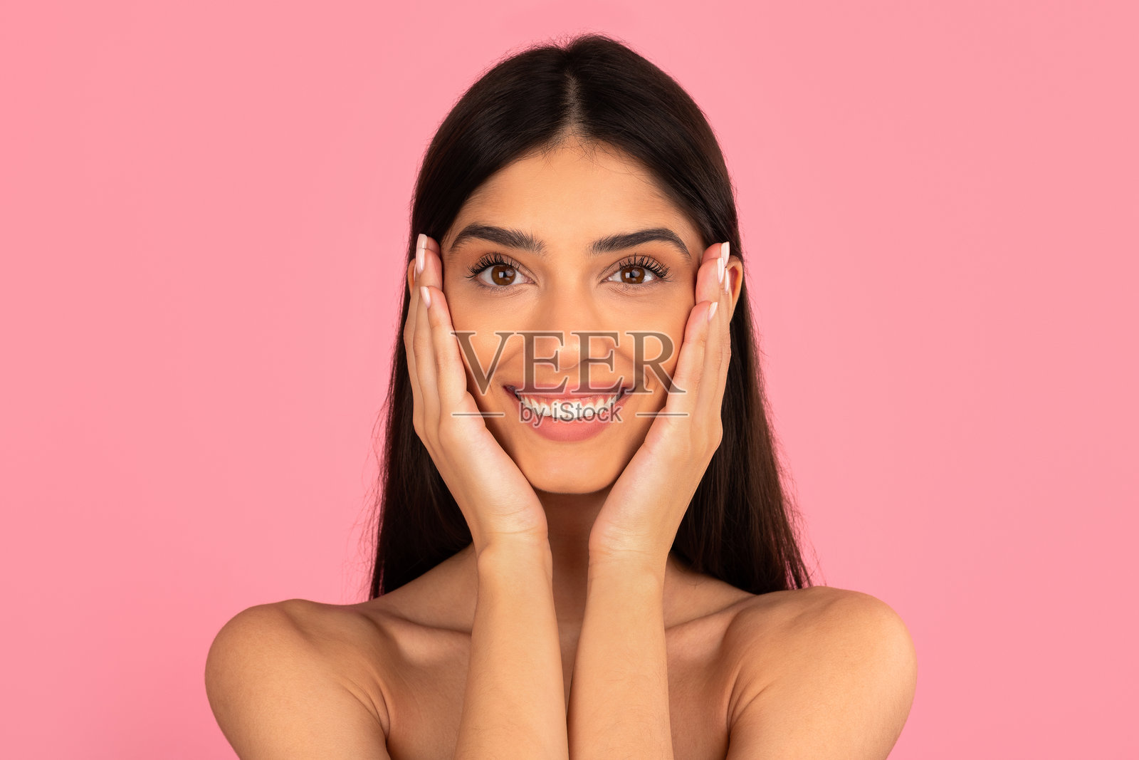 年轻女子开心地双手捧脸，对着粉色背景微笑照片摄影图片