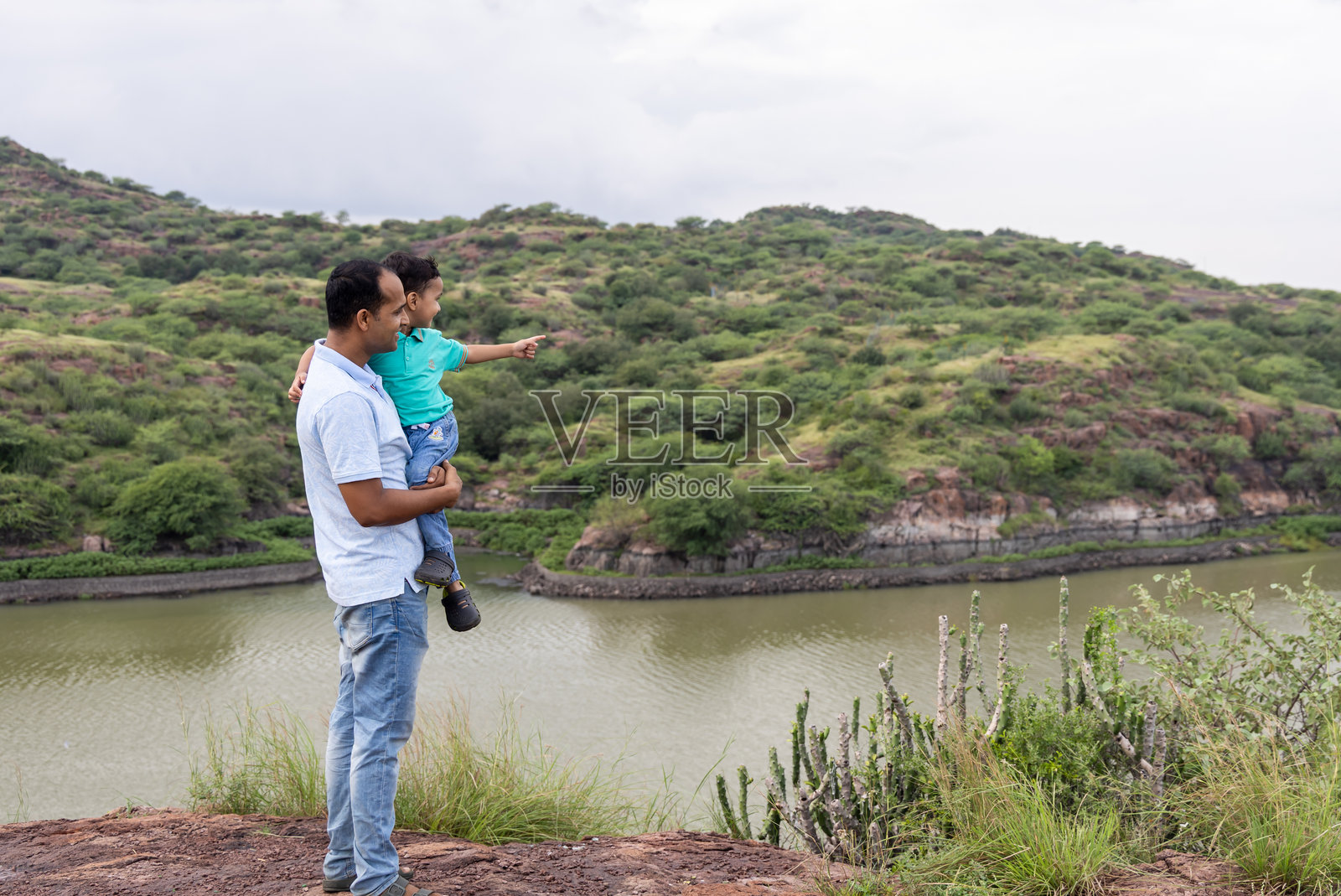 父亲抱着儿子，欣赏着河水和绿色山坡的美景照片摄影图片