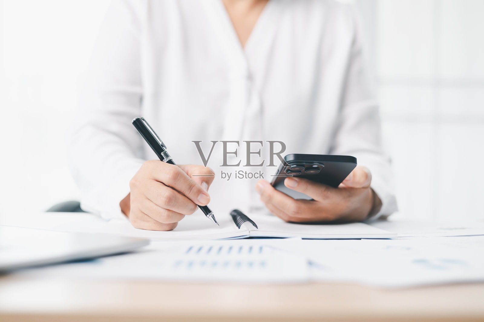 商务规划与生产力概念。一位商人坐在现代办公室办公桌前，一边看手机一边在本子上写笔记，并使用报告和笔记本电脑进行财务分析。照片摄影图片