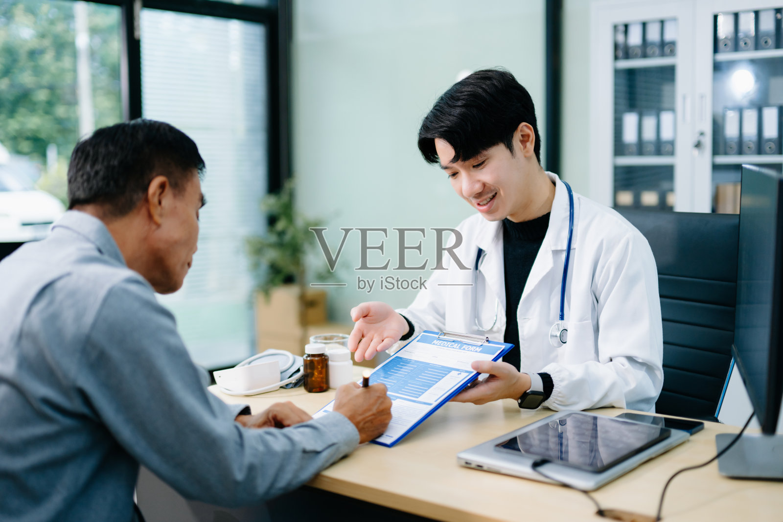 一位亚裔医生在明亮的诊所办公室里，一边咨询病人一边指着一份医疗表格。医疗保健、保险概念照片摄影图片