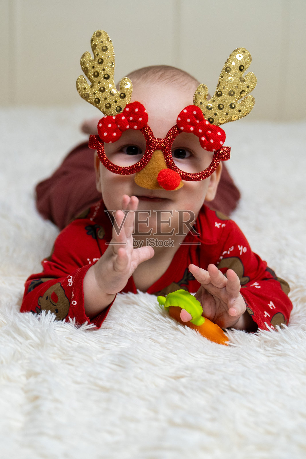 一个穿着红色圣诞睡衣的婴儿，戴着有趣的驯鹿眼镜，眼镜上有鹿角和红鼻子，躺在柔软的白色毯子上，旁边有一个红灯笼。圣诞快乐，新年快乐。照片摄影图片