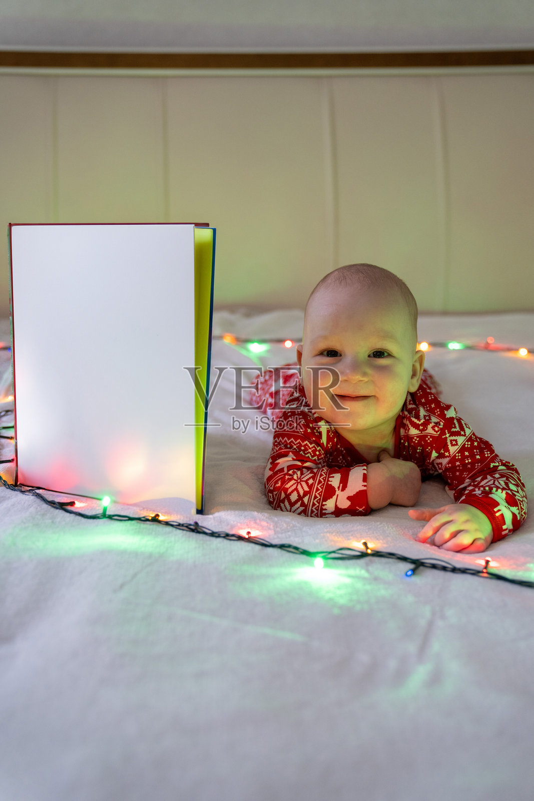 身穿节日红色盛装的微笑婴儿，躺在柔软的白色表面上，周围环绕着闪烁的圣诞串灯。婴儿旁边放着一本竖立的空白书本或浅色面板。照片摄影图片