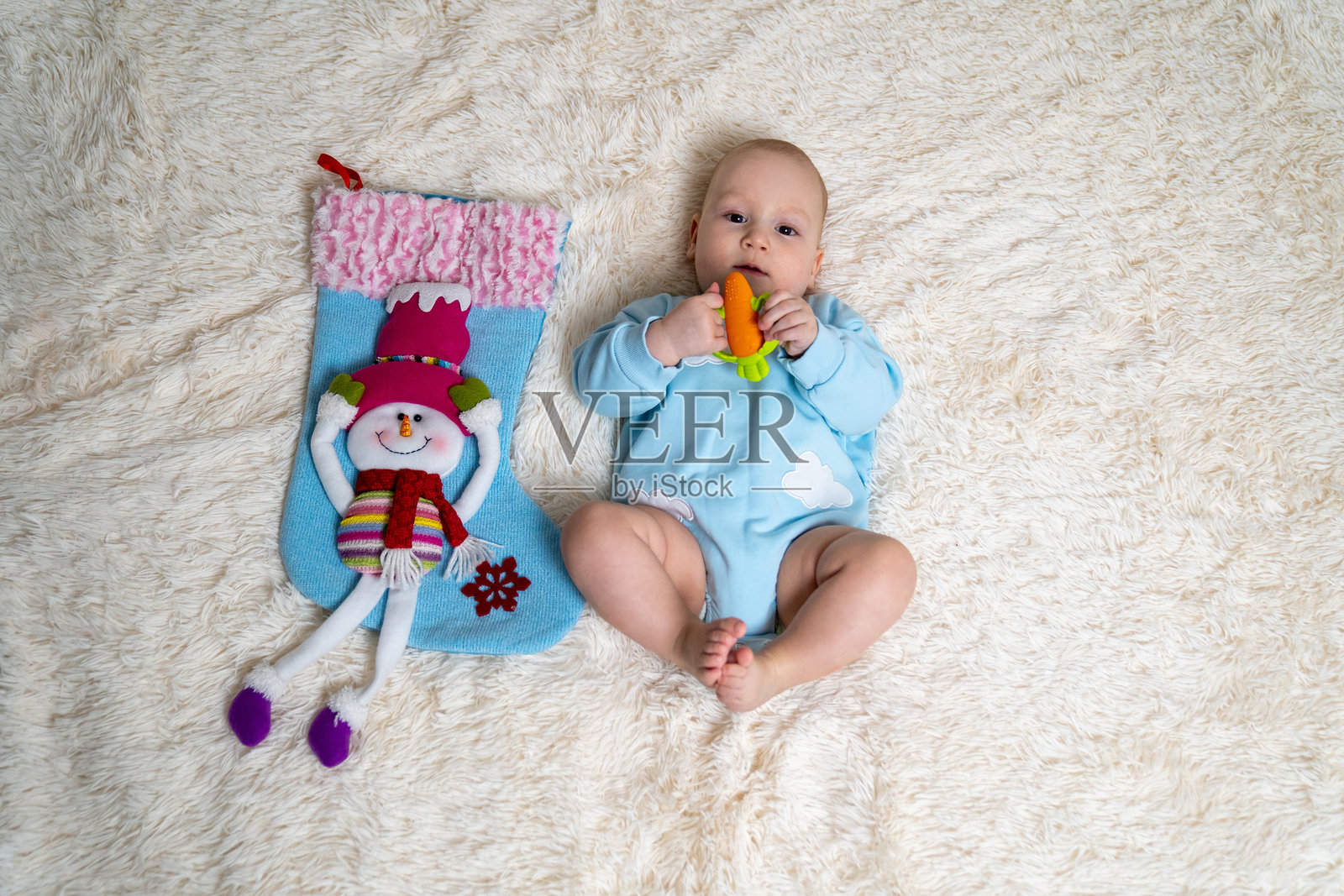 一个穿着浅蓝色连体衣的可爱婴儿，躺在柔软的米色毯子上，手里拿着一个色彩鲜艳的牙胶玩具。婴儿旁边是一个大号的蓝色圣诞袜，上面装饰着一个穿着节日冬装的快乐雪人。照片摄影图片