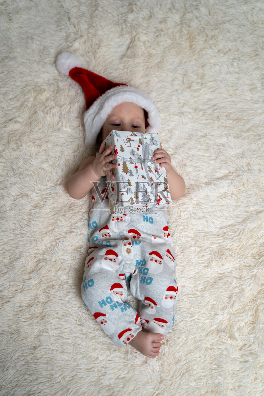 可爱的婴儿戴着圣诞老人帽和节日“Ho Ho Ho”圣诞睡衣，躺在柔软蓬松的毯子上，手里拿着一份包装好的节日礼物。照片摄影图片