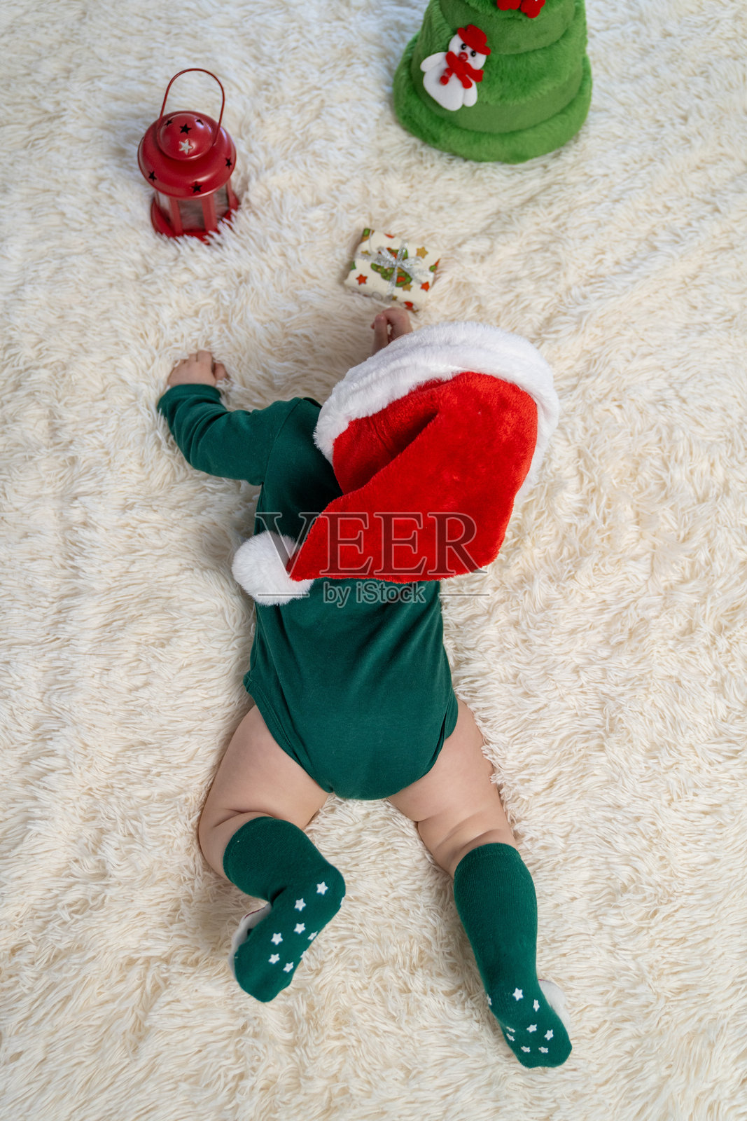 穿着圣诞帽和绿色圣诞服装的婴儿的背面，躺在一个毛茸茸的地毯上，旁边有节日装饰品——一个小灯笼、一个包装好的礼物和一个节日装饰品。小精灵婴儿照片摄影图片