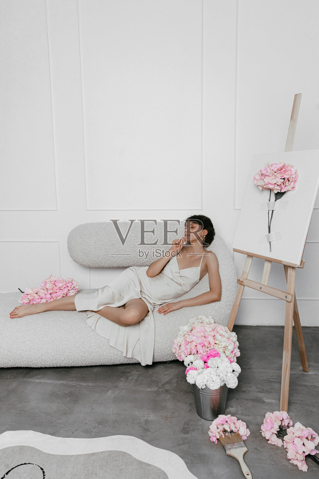 身着浅色连衣裙的女子正舒适地躺在沙发上，身处一个充满粉色和白色鲜花的极简主义艺术工作室，她正全神贯注地在画架上创作一幅花卉画作。照片摄影图片