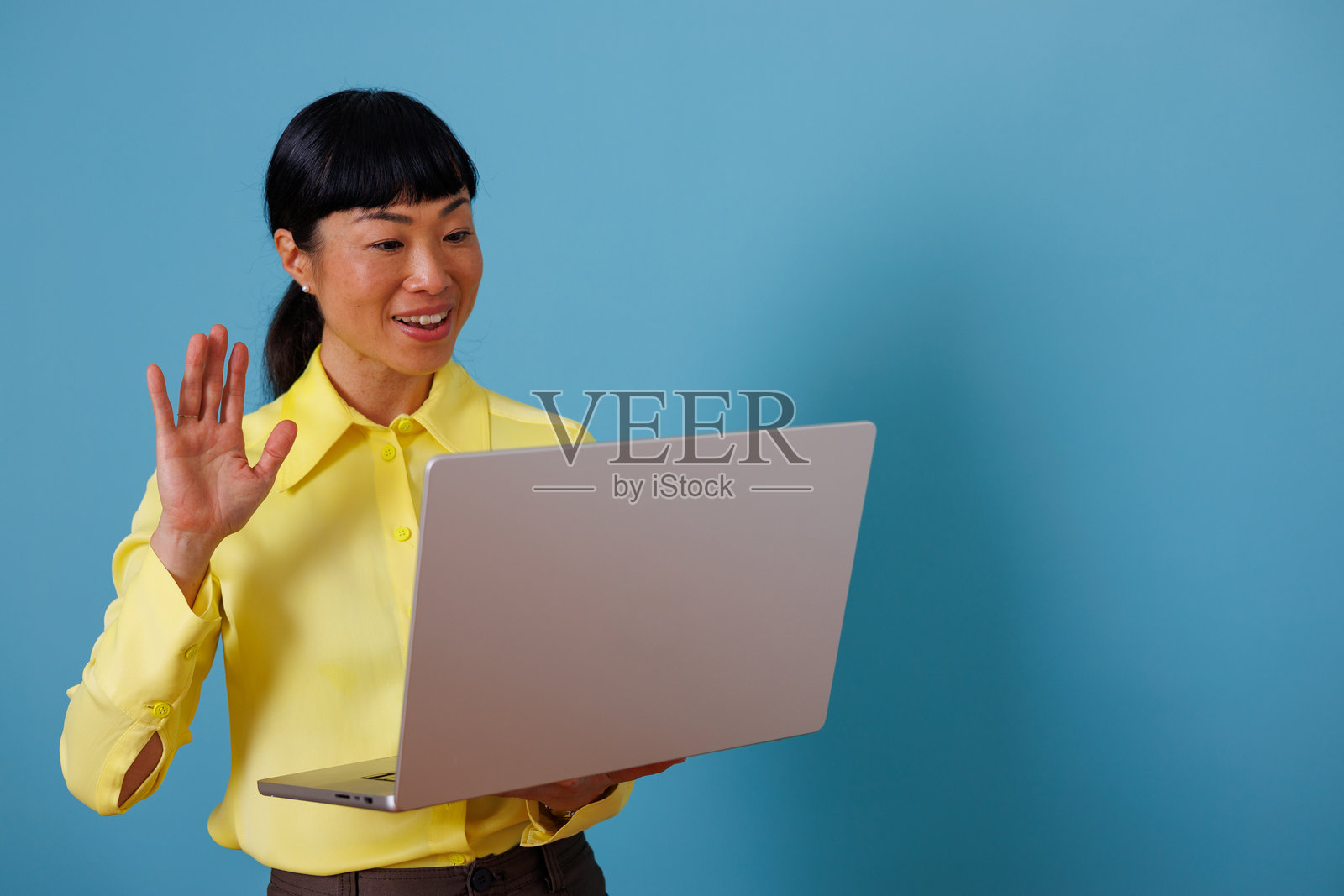 一位亚洲女性正在使用笔记本电脑进行视频交流，在在线会议或虚拟会议中挥手，背景为蓝色照片摄影图片