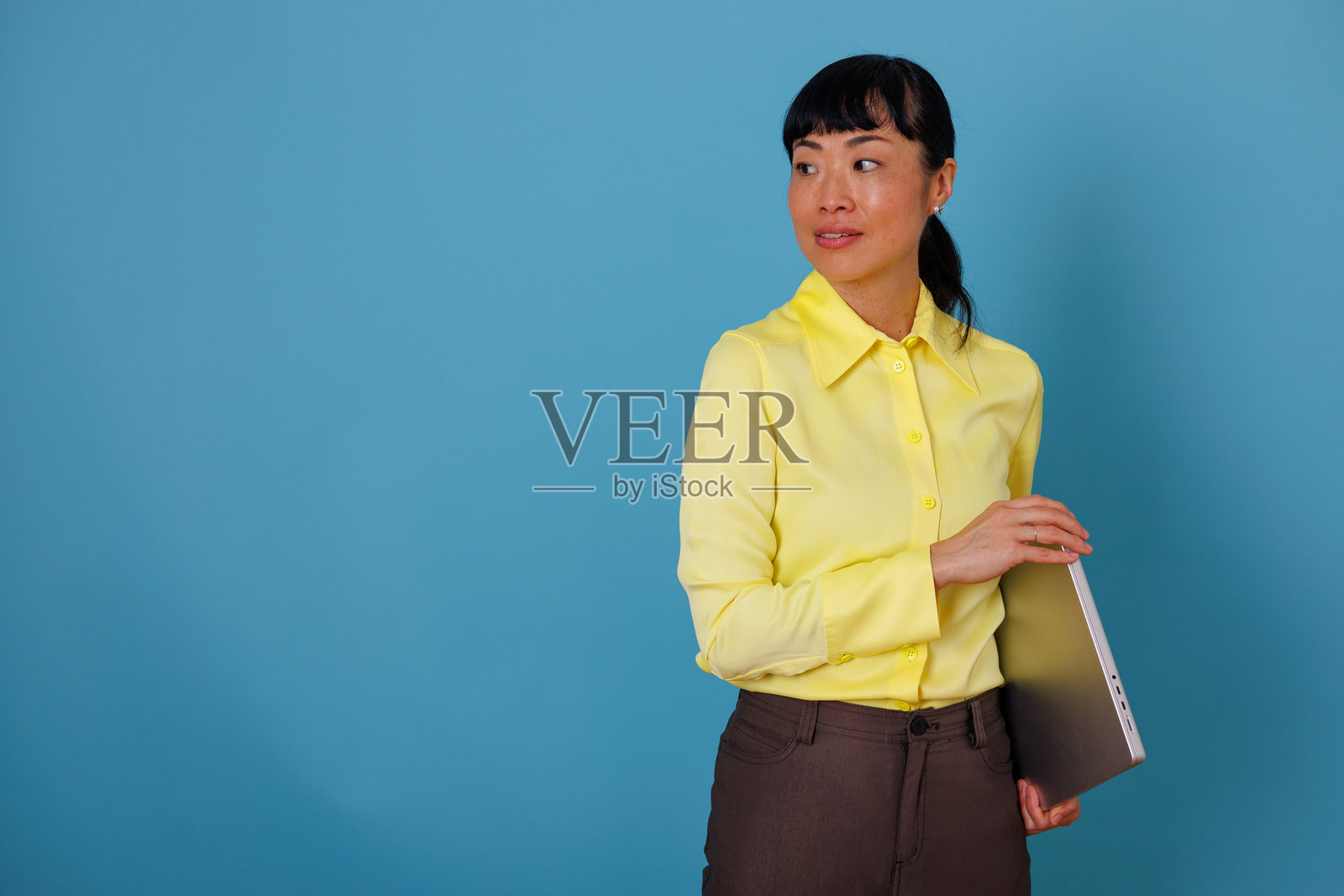 一位亚洲女商人手持笔记本电脑，背对蓝背景，望向别处，留有复制空间。照片摄影图片