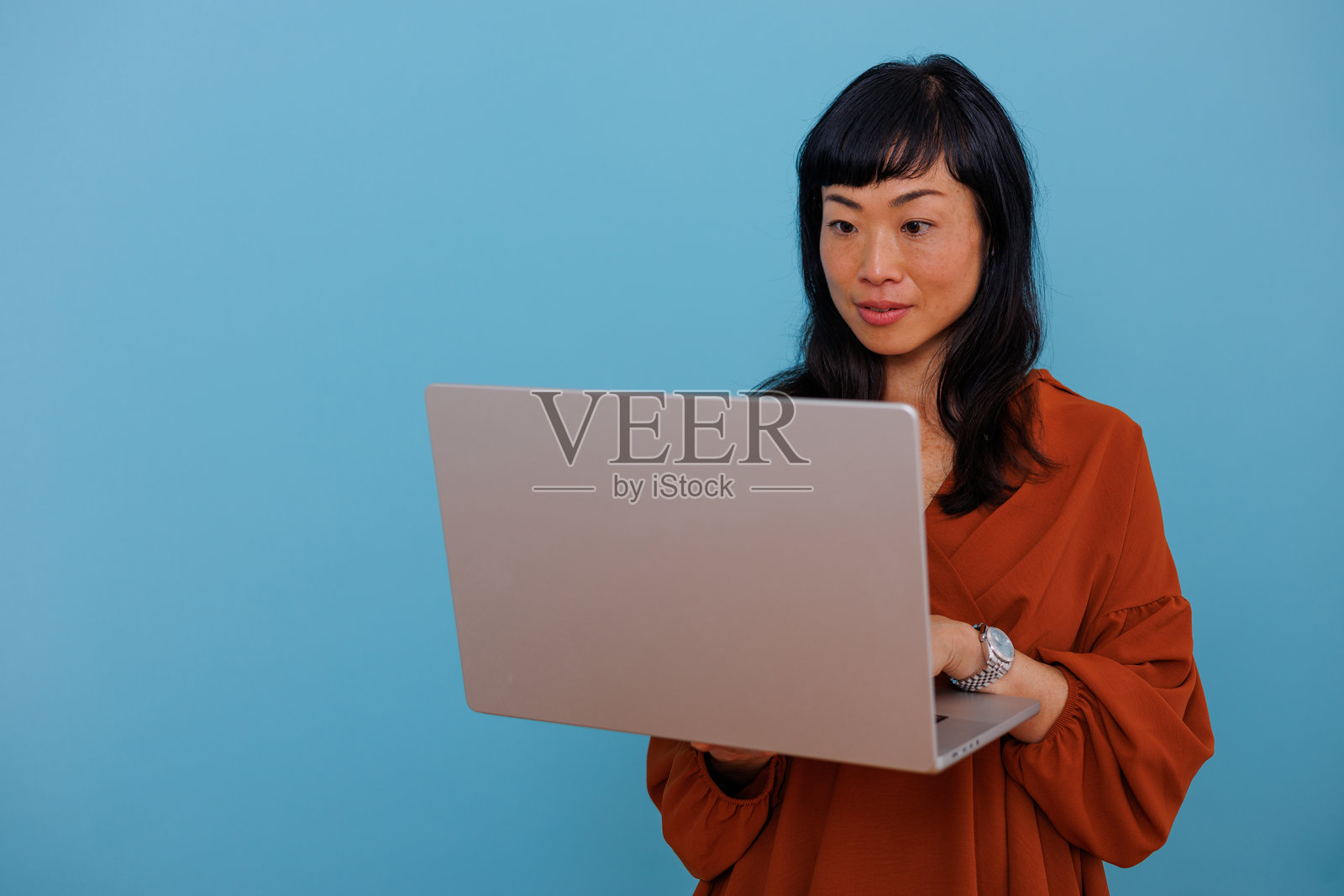 专业亚洲女性在蓝色背景下使用笔记本电脑工作，专注于屏幕，体现数字化商业理念照片摄影图片