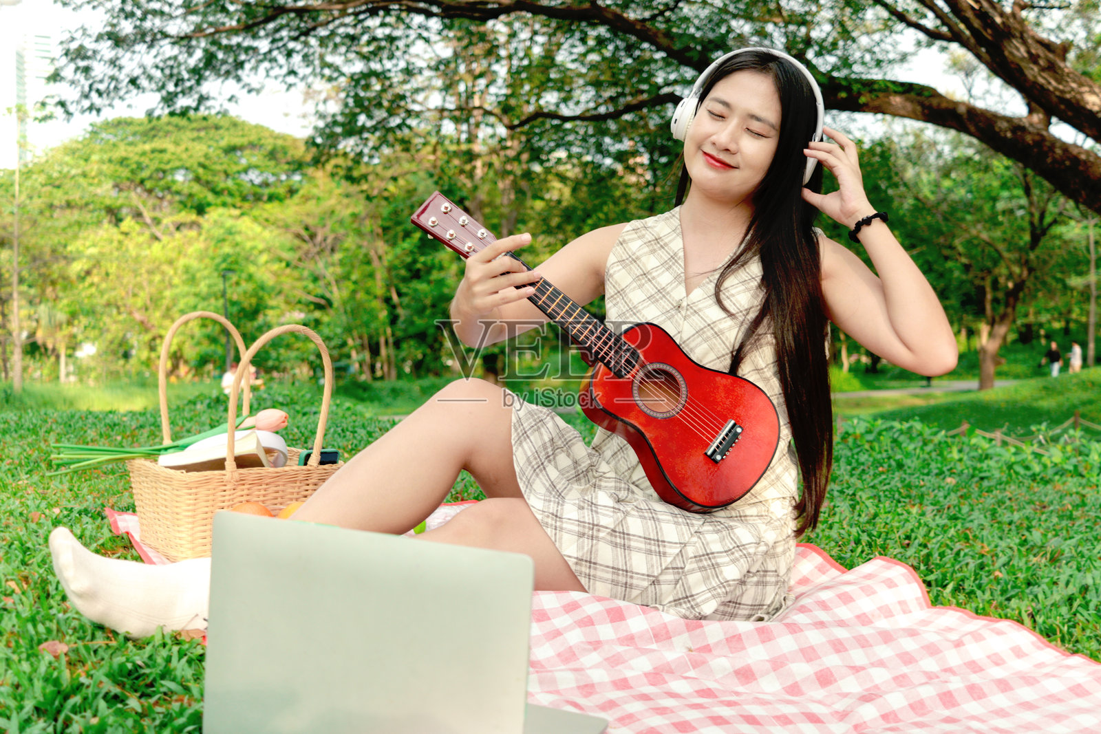 微笑的亚洲美女在公园野餐时弹奏尤克里里。年轻女子在绿色花园的野餐中放松身心。快乐的女性在大自然中度过户外时光。照片摄影图片