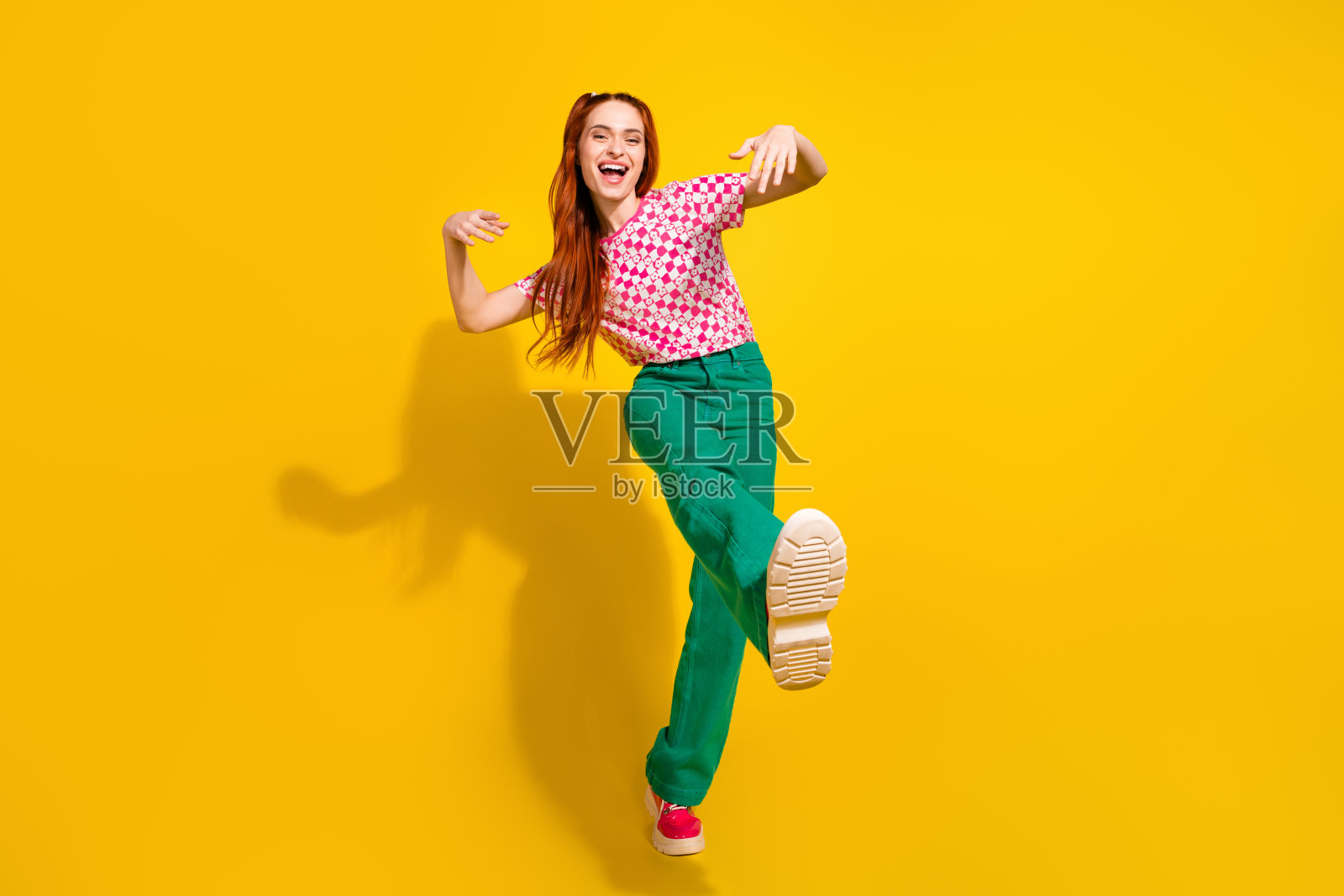 穿着格纹粉色衬衫和绿色裤子的快乐小女孩，在明黄色的背景前跳舞，展现时尚活力照片摄影图片
