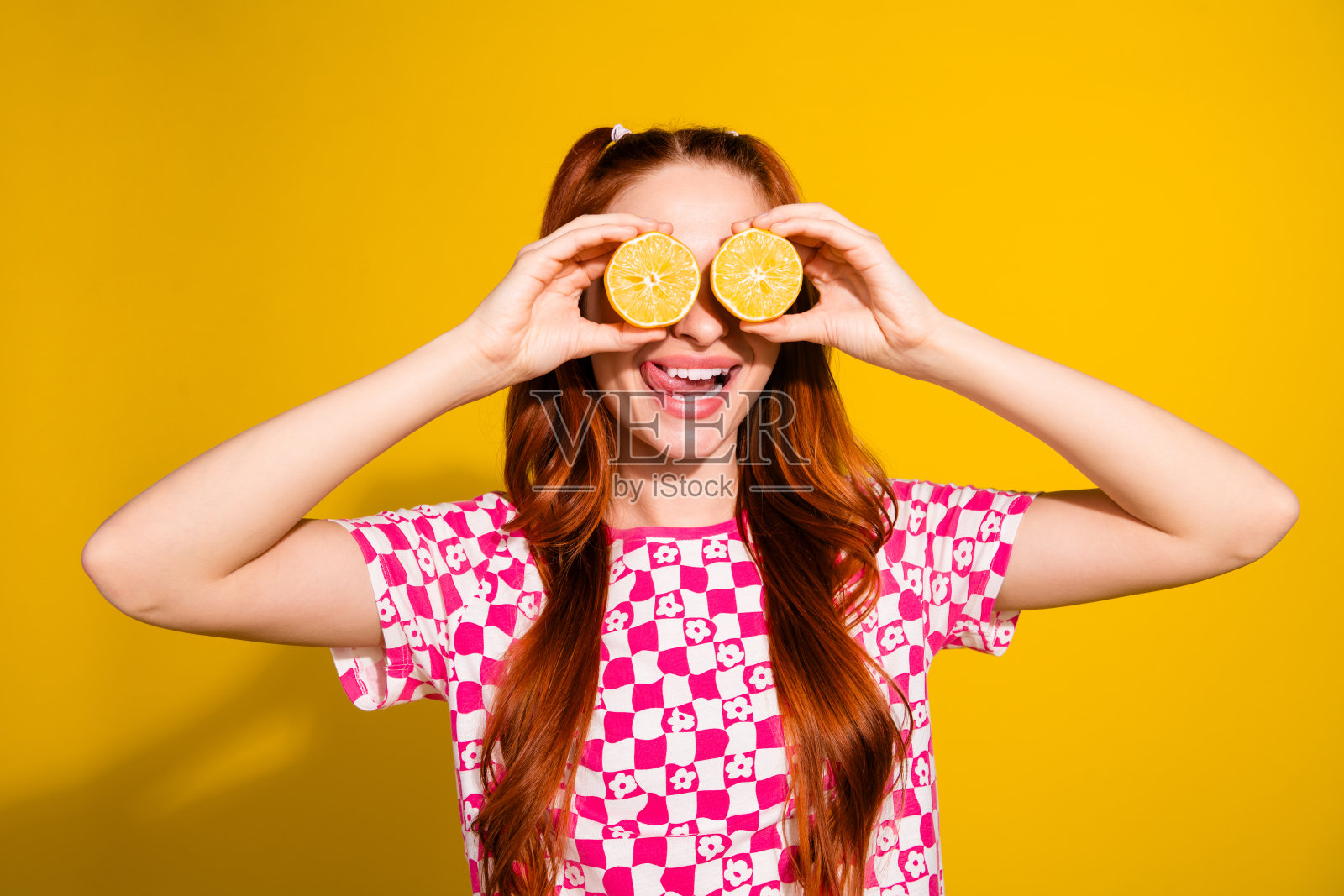 一位红发年轻女子身穿粉色格纹衬衫，将橙子片放在眼前，背景为明亮的黄色。照片摄影图片