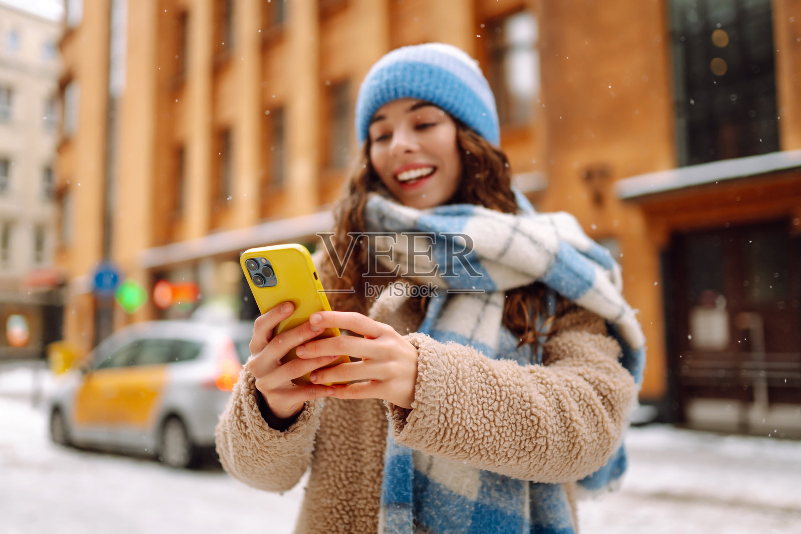 穿着暖和的年轻女子在雪天的城市街道上近距离使用手机。照片摄影图片