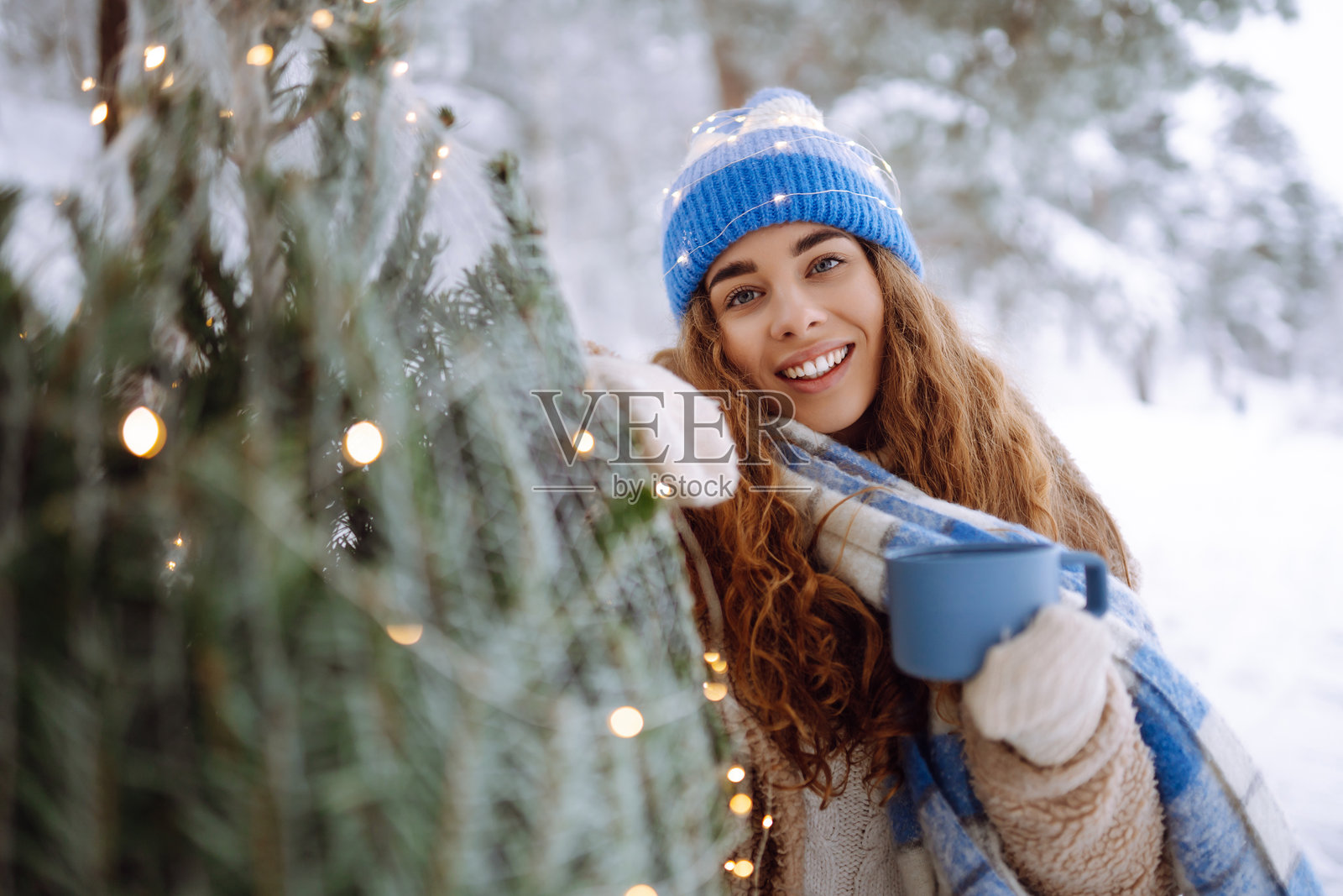 一位美丽的女子在冬季森林中，靠近一棵绿色的圣诞树，她正享受着蓝色保温瓶里的热茶。照片摄影图片
