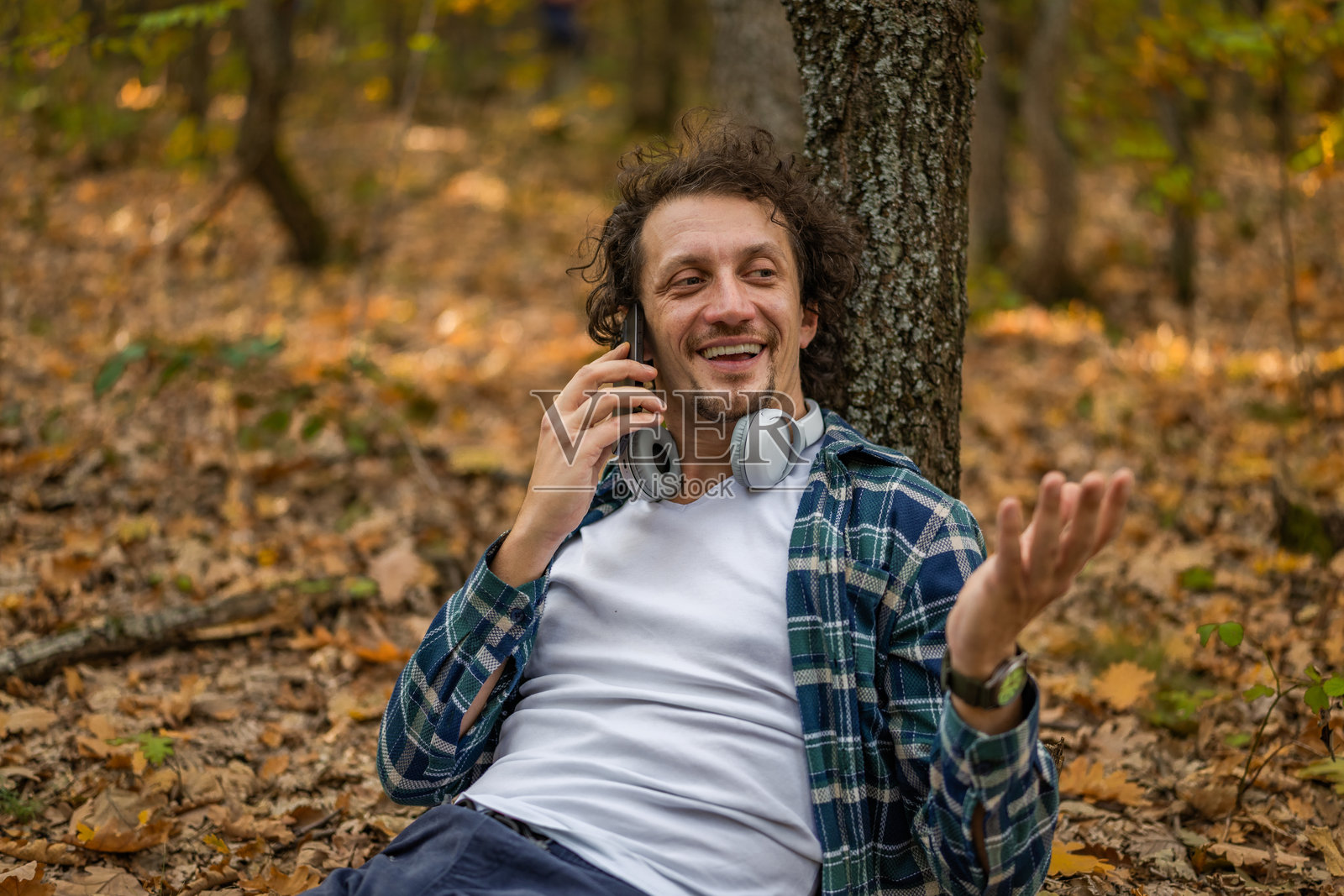 一个卷发男子在秋日里，倚靠在树干上，一边看着手机一边在大自然中休憩。照片摄影图片