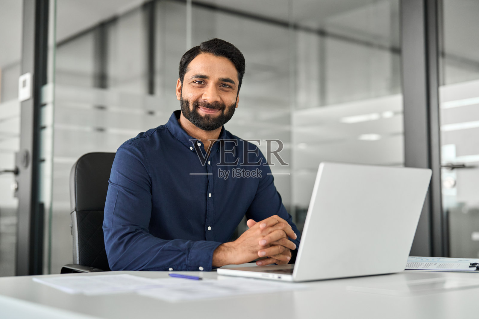 一位自信微笑的印度商人坐在办公桌前使用笔记本电脑。肖像照片摄影图片