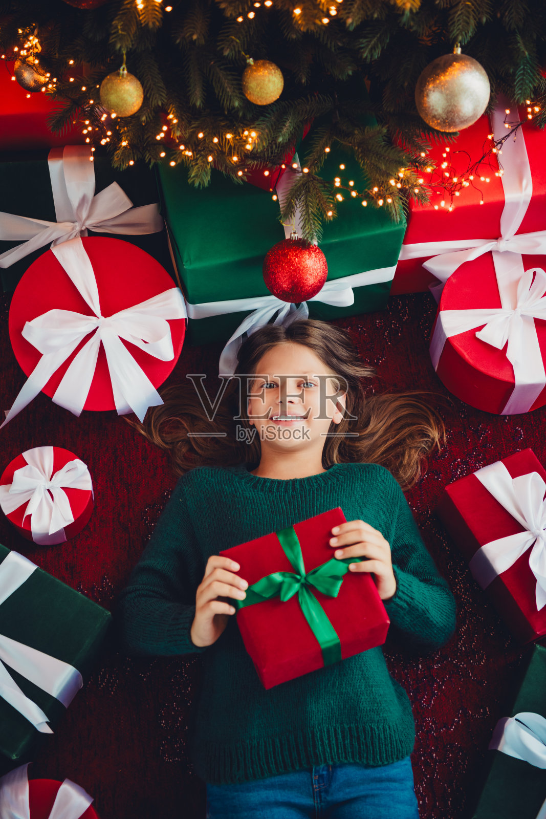 一个穿着绿色毛衣的小女孩，在闪烁的圣诞树下抱着一个红色礼物，周围是五颜六色的礼品和温馨的家居装饰，这是一个有趣的节日之夜的梦想。照片摄影图片