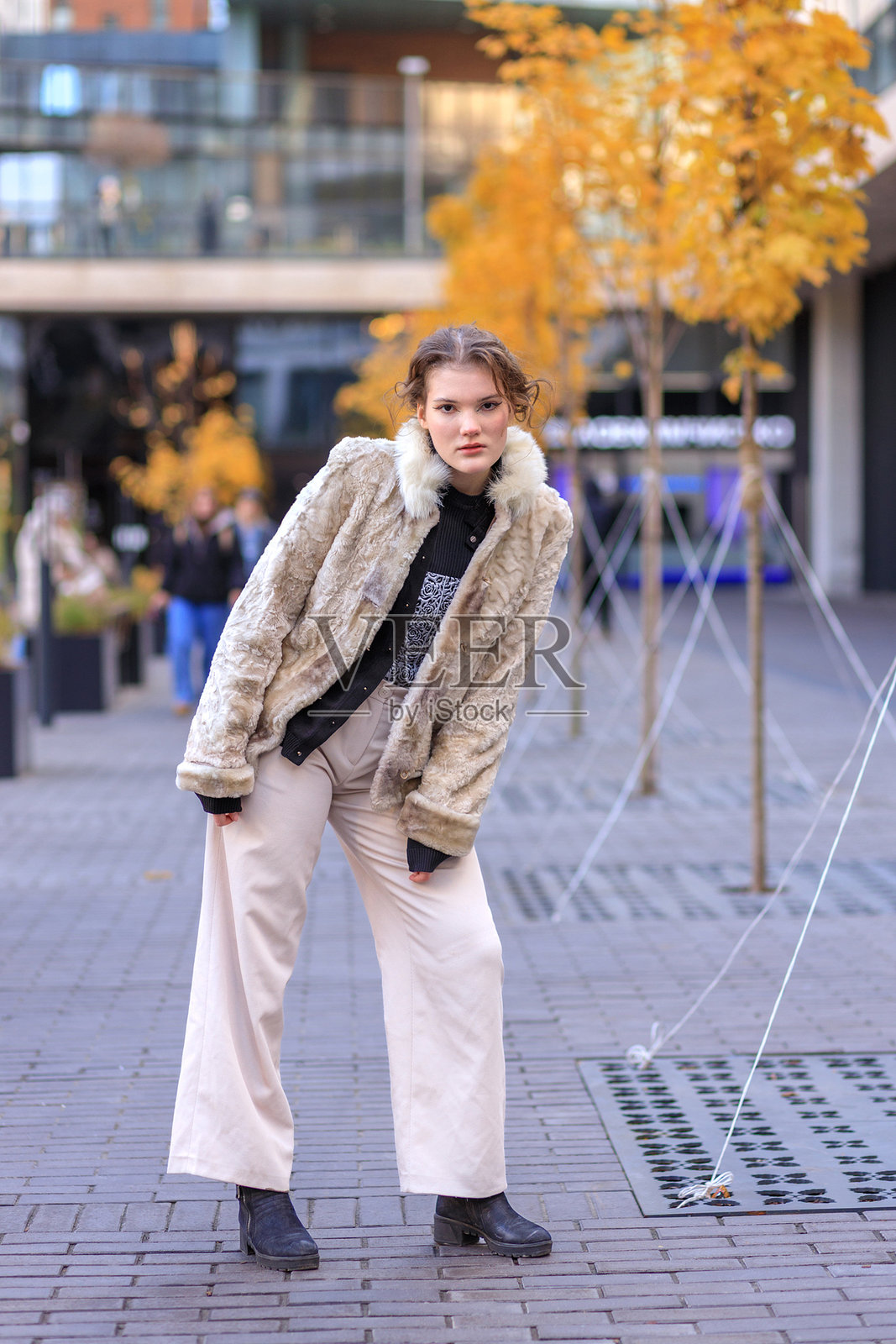 穿着浅色暖色调衣服的年轻棕发女子走在秋日城市街道上。照片摄影图片