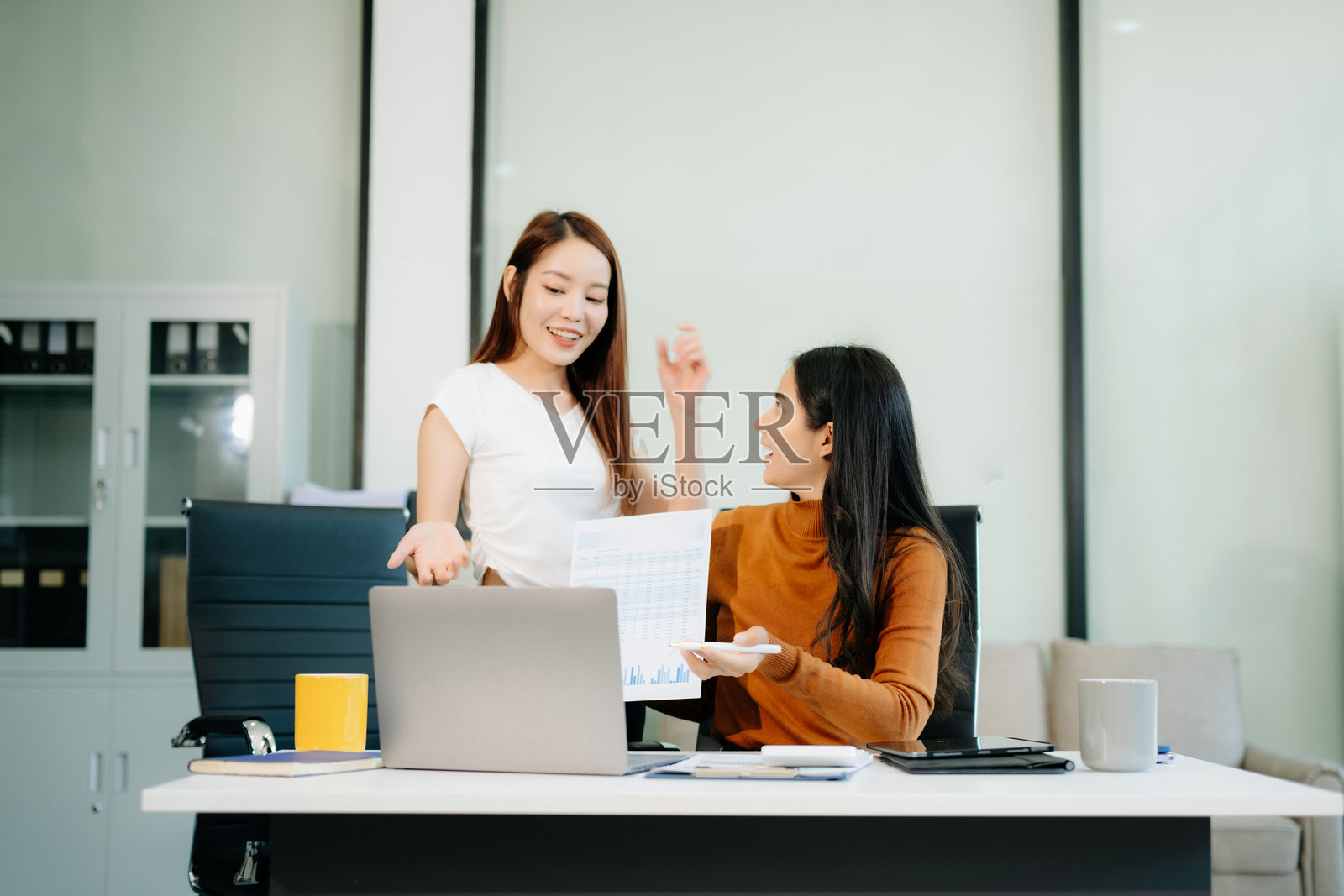 两位亚洲女性的商务肖像，她们面带笑容，愉快地交谈，手中拿着平板电脑，举起双手，表示赞同，准备好迎接新项目。照片摄影图片