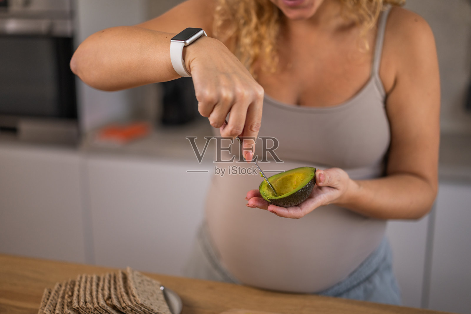 一位卷发孕妇在准备健康的早餐餐点时，挖出牛油果的果肉。照片摄影图片