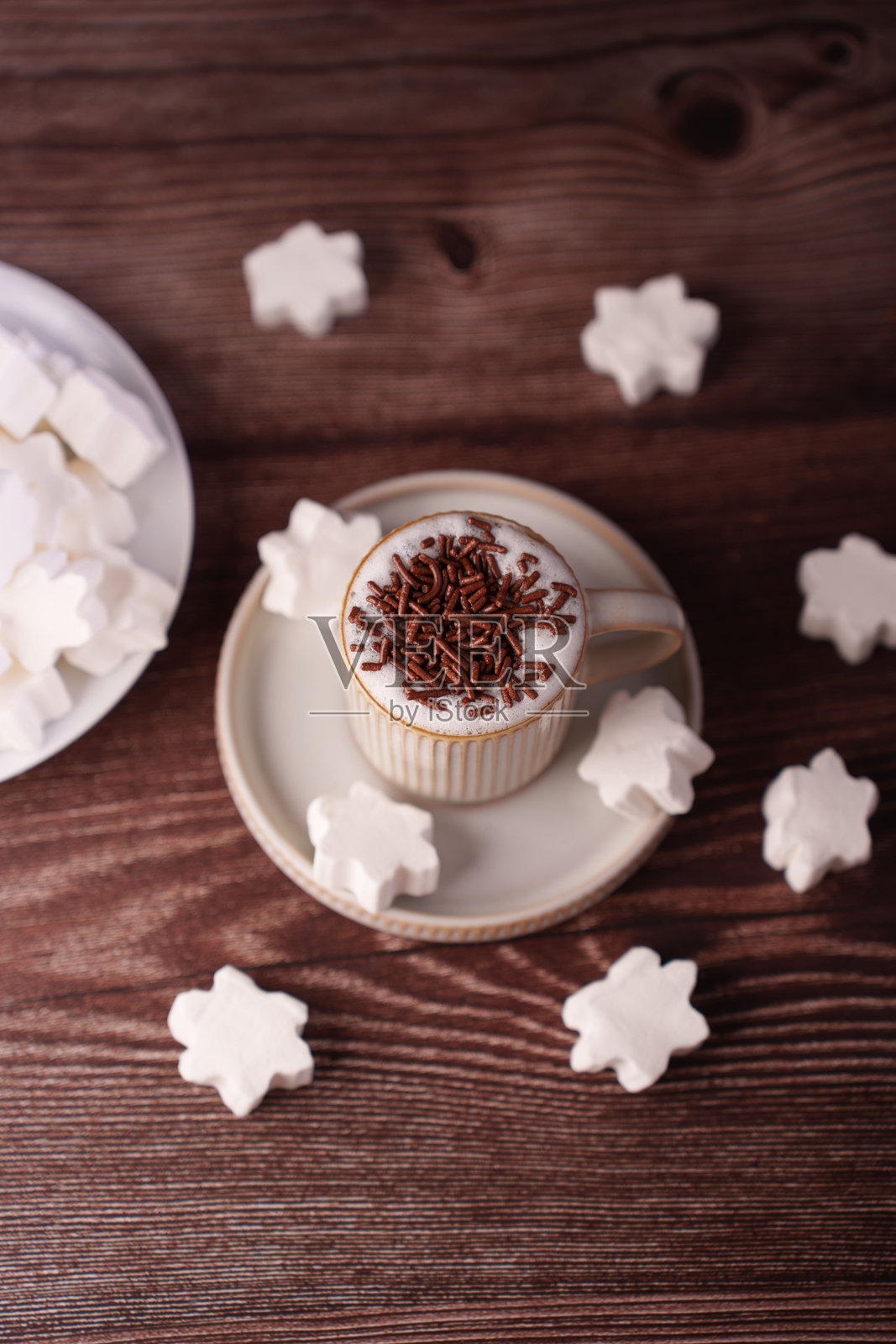 一杯浓郁的卡布奇诺咖啡，上面撒着巧克力碎，周围环绕着雪花形状的棉花糖，摆放在温暖的木质表面上，营造出温馨、艺术和美食的氛围。照片摄影图片