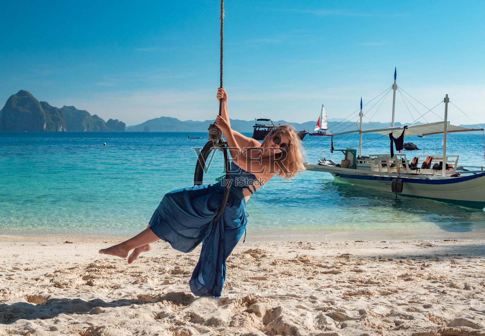 一位长发金发女子身穿蓝色连衣裙，在清澈碧绿的水面上荡着轮胎秋千。照片摄影图片