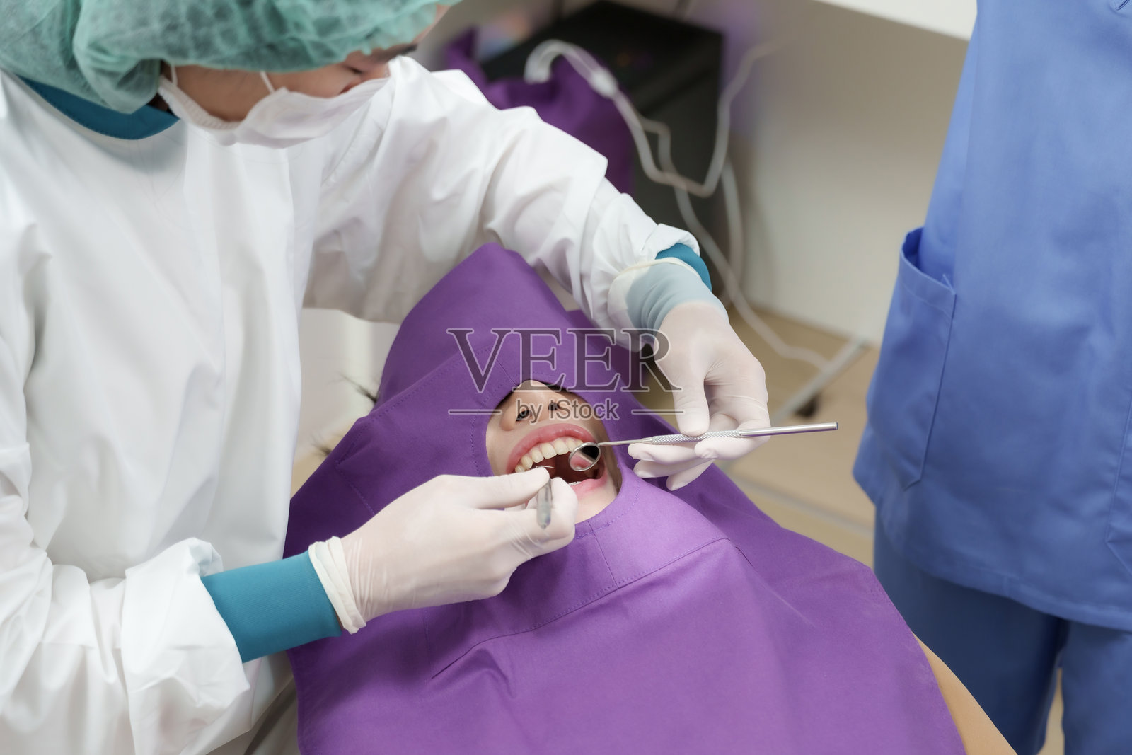 身穿白色制服的亚洲女牙医使用牙科镜和探针，在专业的牙科诊所内检查被紫色围布盖住的成年女患者的牙齿，进行口腔检查照片摄影图片